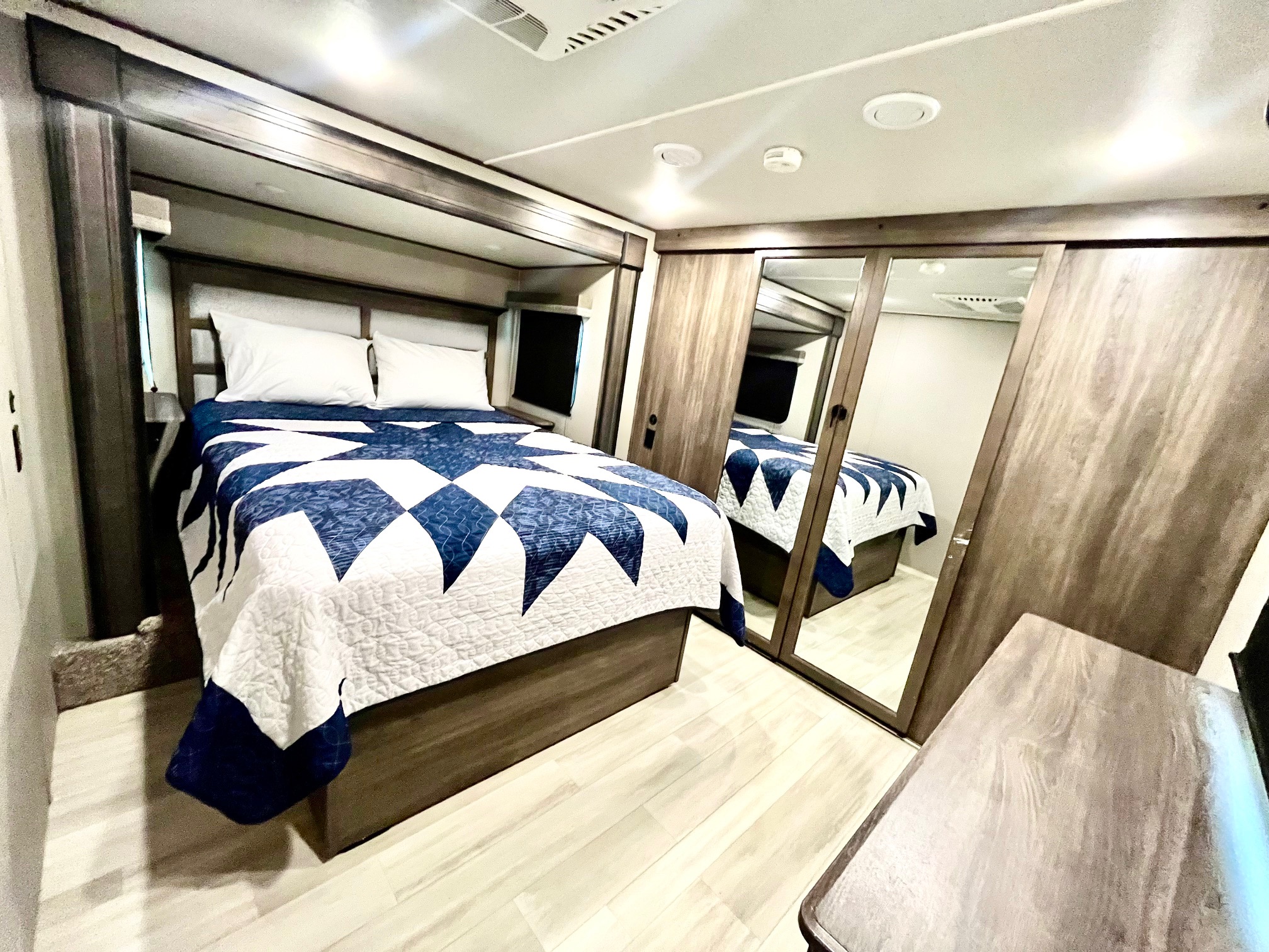 Modern RV rental bedroom featuring a cozy bed with a blue and white quilt, mirrored closet doors, and light wood-look flooring.. Grand Design Solitude 2021