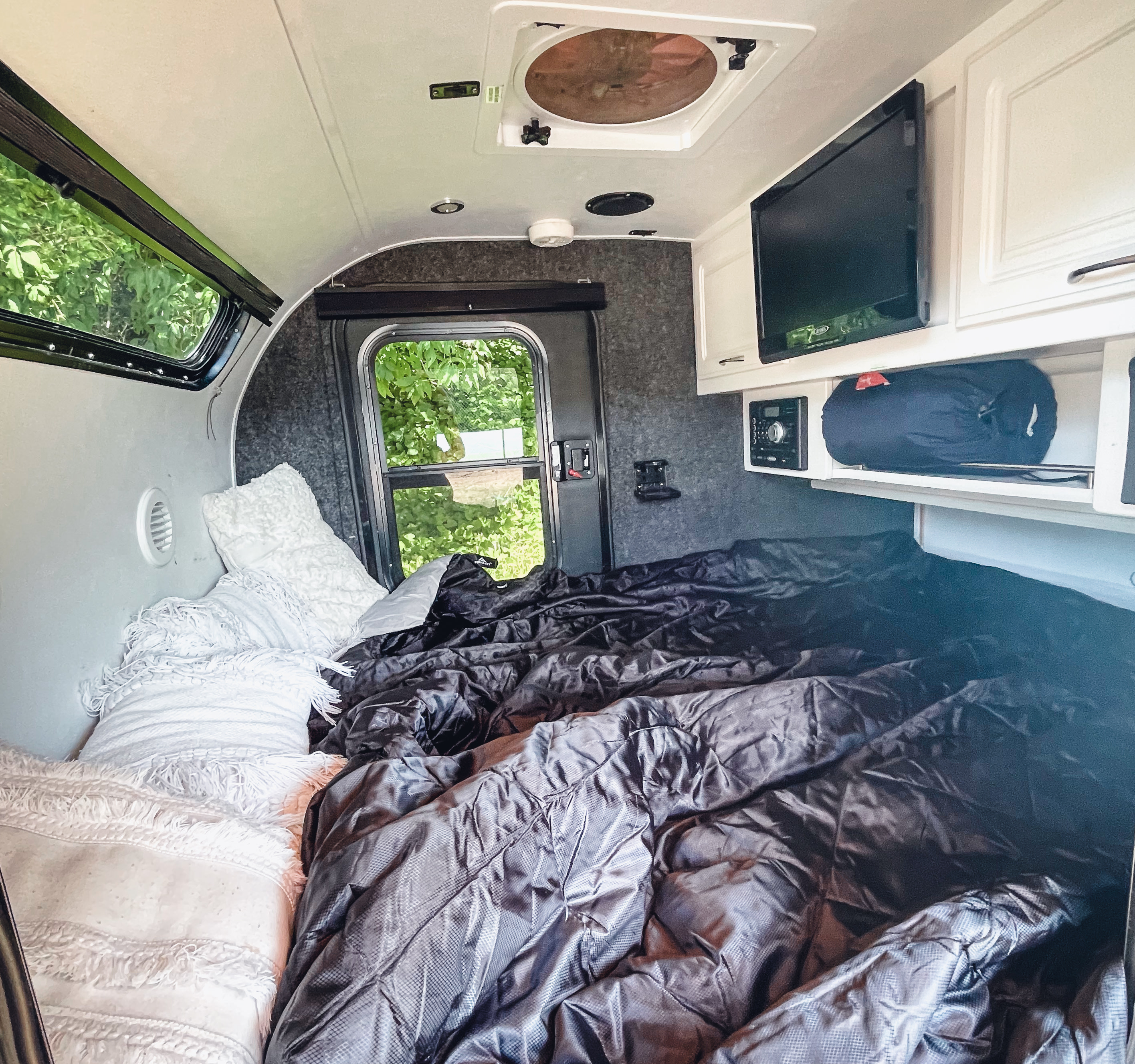 Inside a compact RV rental, showing a neatly made dark bed, TV, cabinets, and lush green outdoor views.. Little Guy Little Guy Trailer 2016