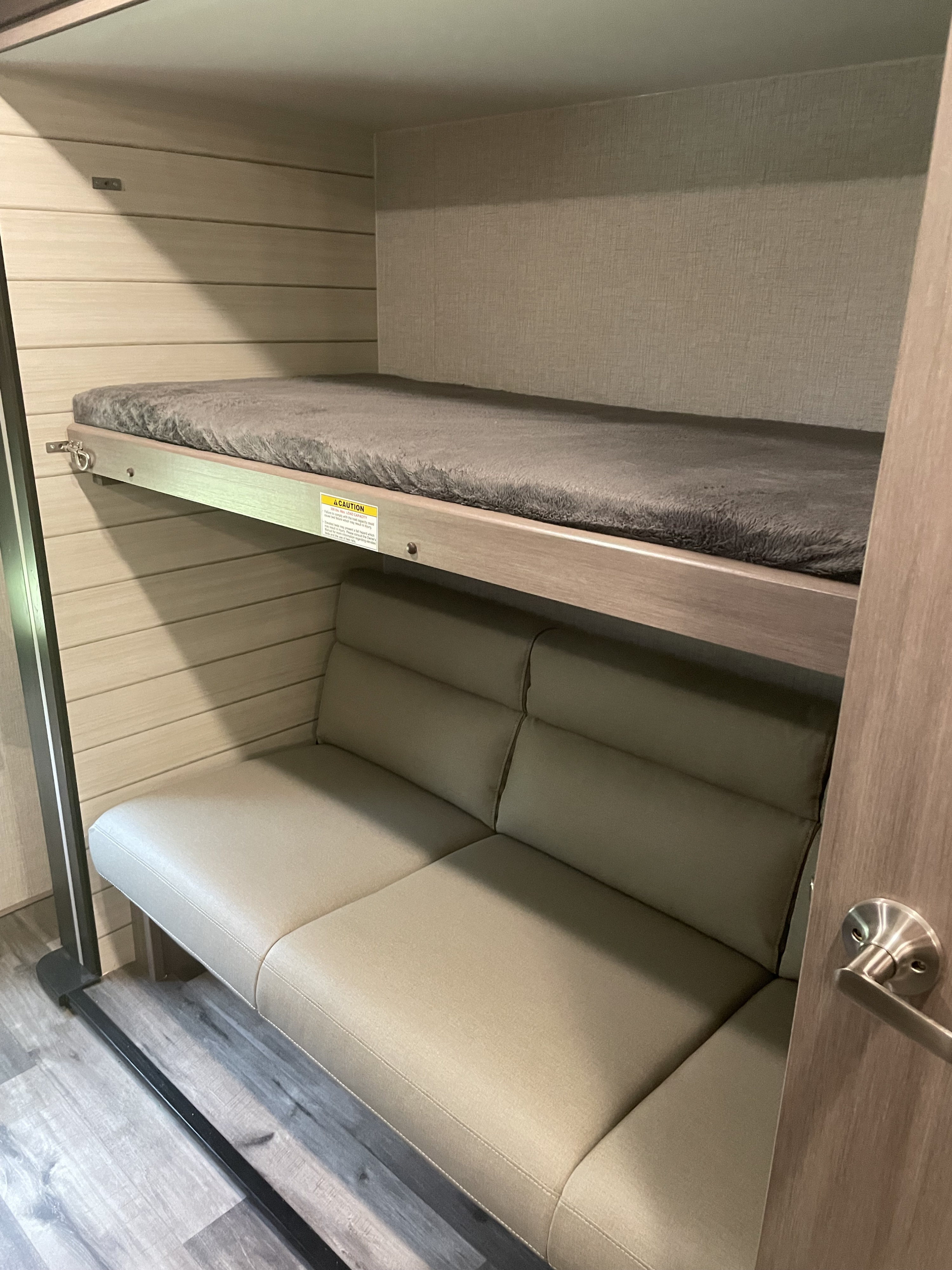 RV rental interior: A compact bunk bed with a gray fuzzy mattress is set above a light-toned sofa/seating area.. Crossroads RV Sunset Trail Super Lite 2023