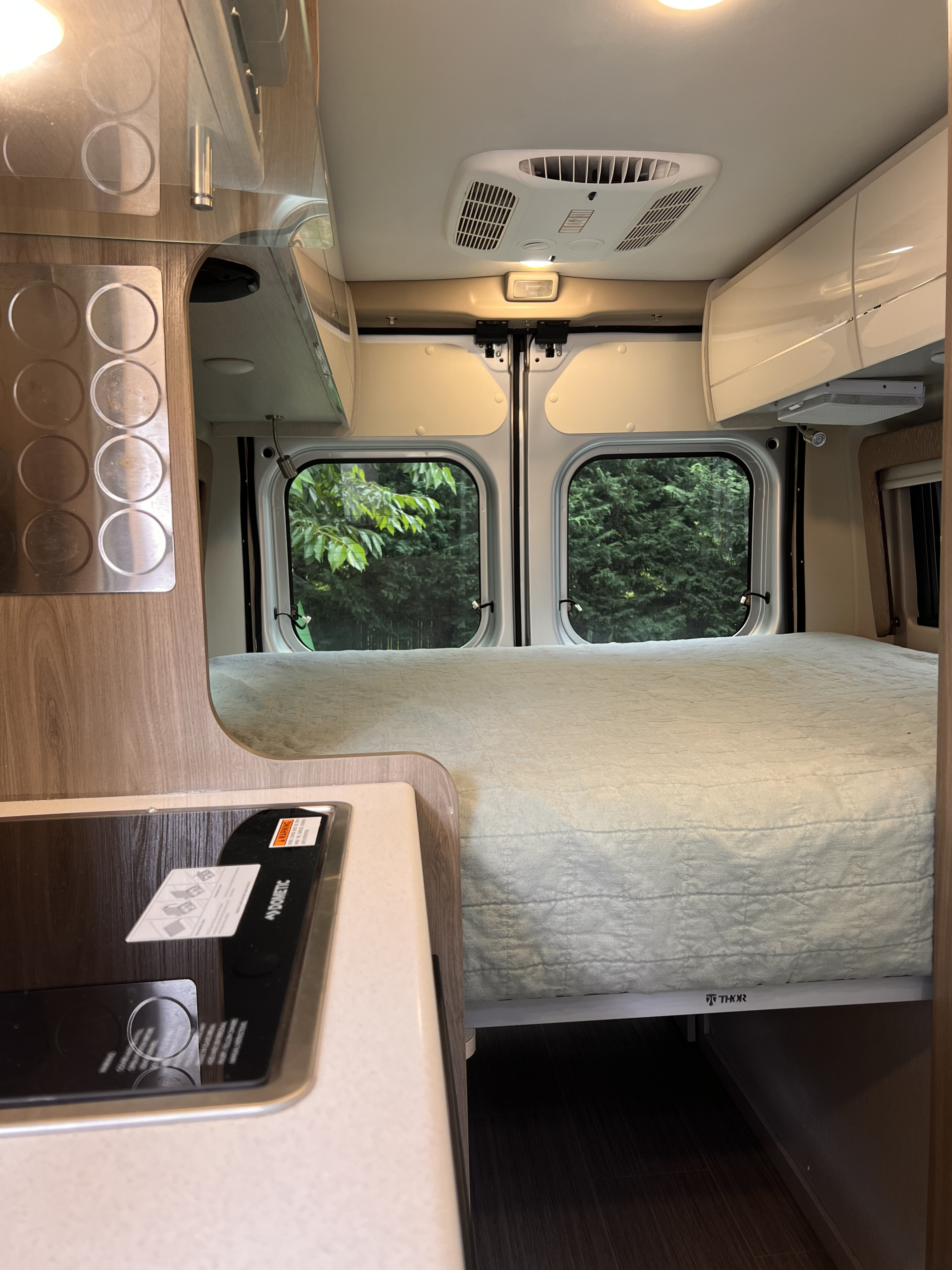 Cozy rv rental interior: view of the comfy bed, kitchen area with Dometic cooktop, and large windows with nature views.. Thor Tellaro 2022