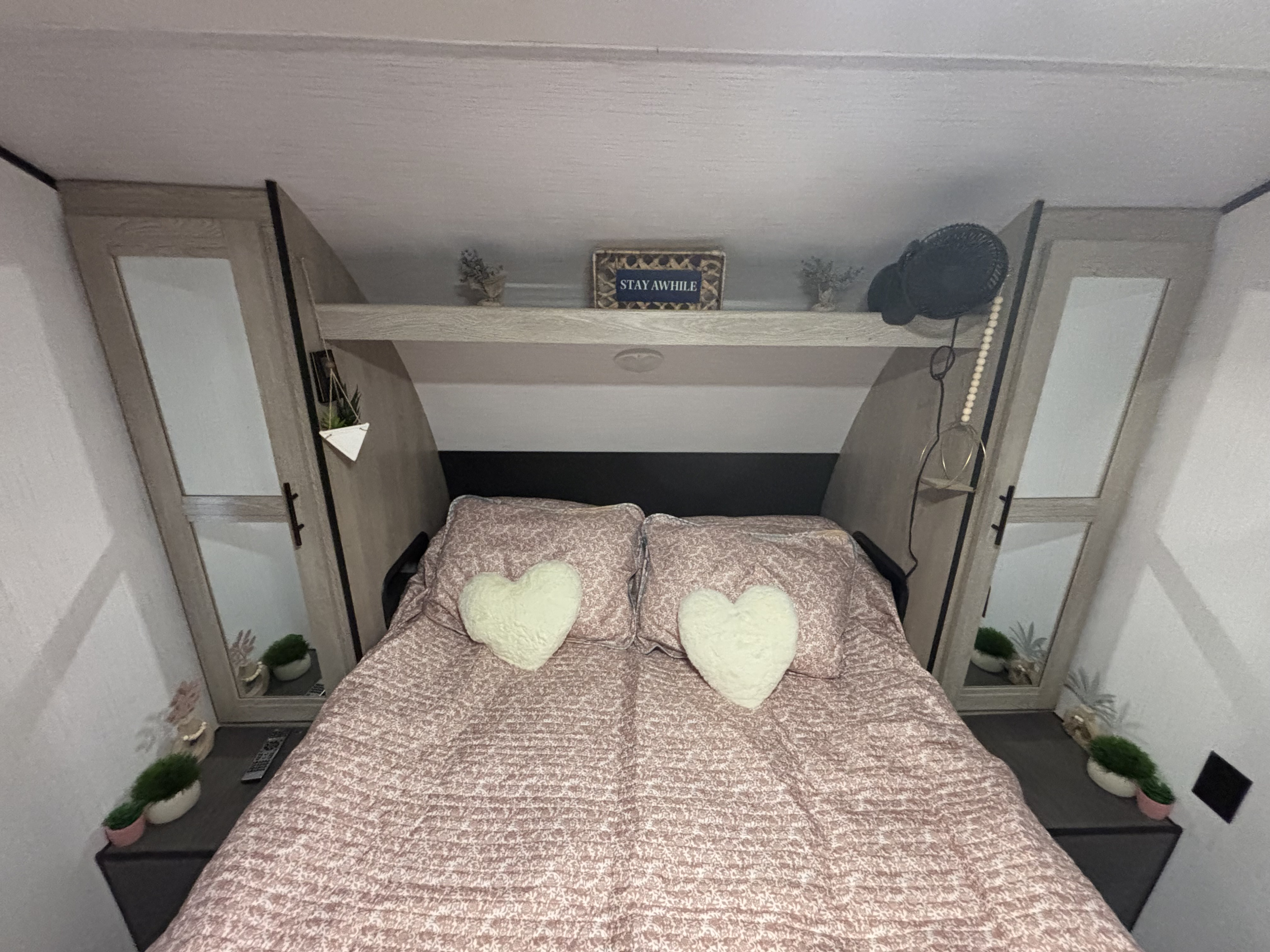 Relax in this charming rv rental bedroom! Features a comfy bed with heart pillows, organized overhead storage, and thoughtful decor.. Heartland RVs Prowler 2021