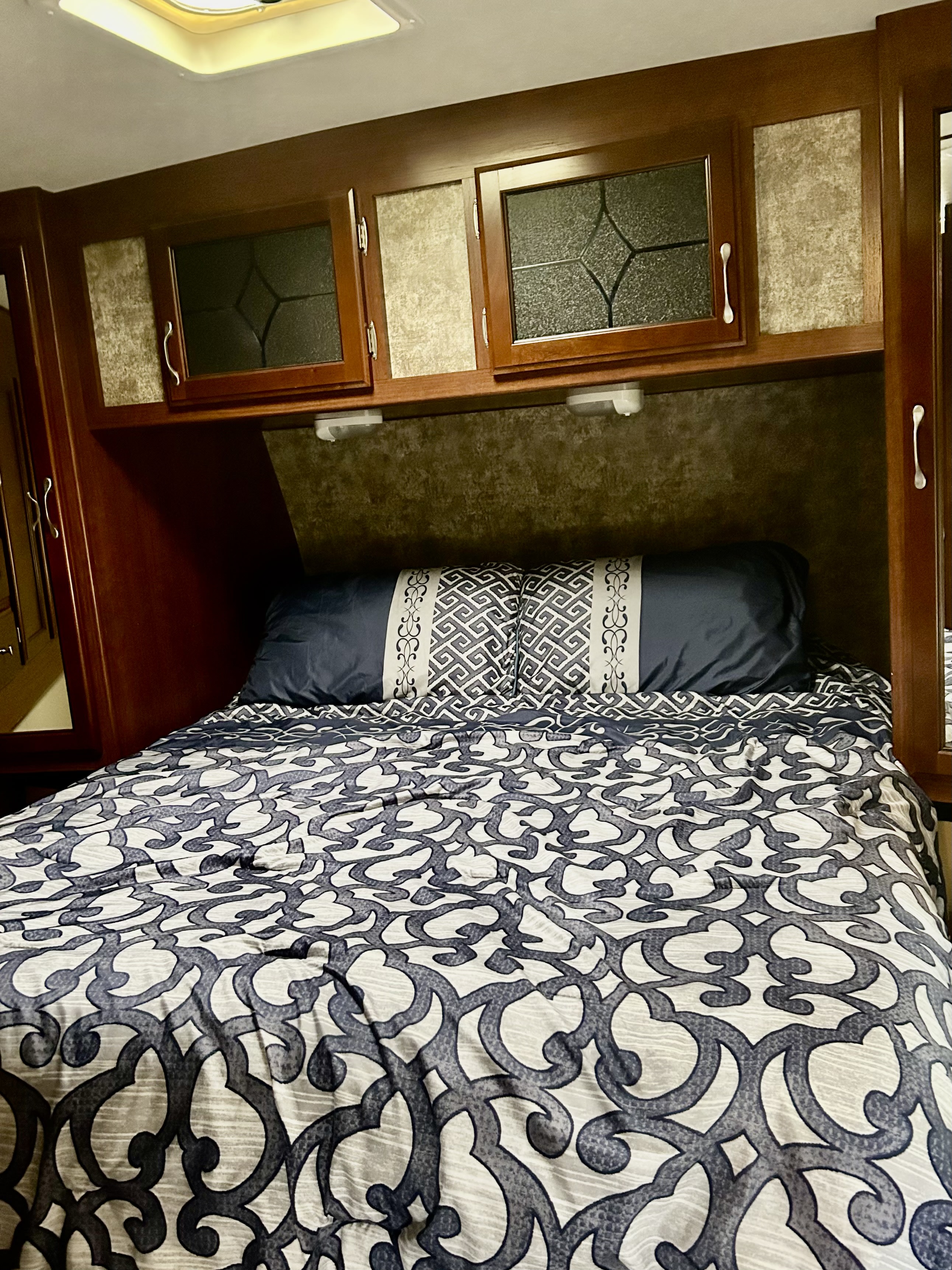 Cozy RV rental bedroom featuring a patterned bedspread, dark wood overhead cabinets with glass inserts, and a ceiling light.. Evergreen Sunvalley 2015