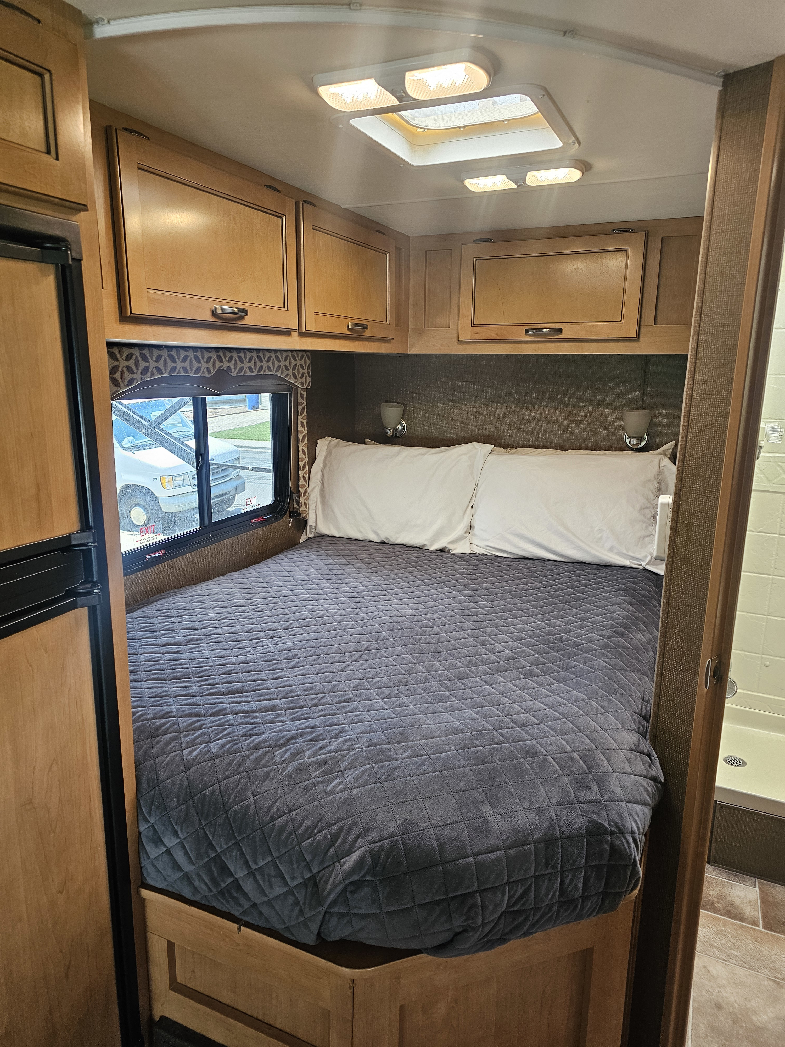 Cozy RV rental bedroom featuring a large bed with white pillows, overhead storage cabinets, a window, and a glimpse of the shower.. Thor Four Winds 2016