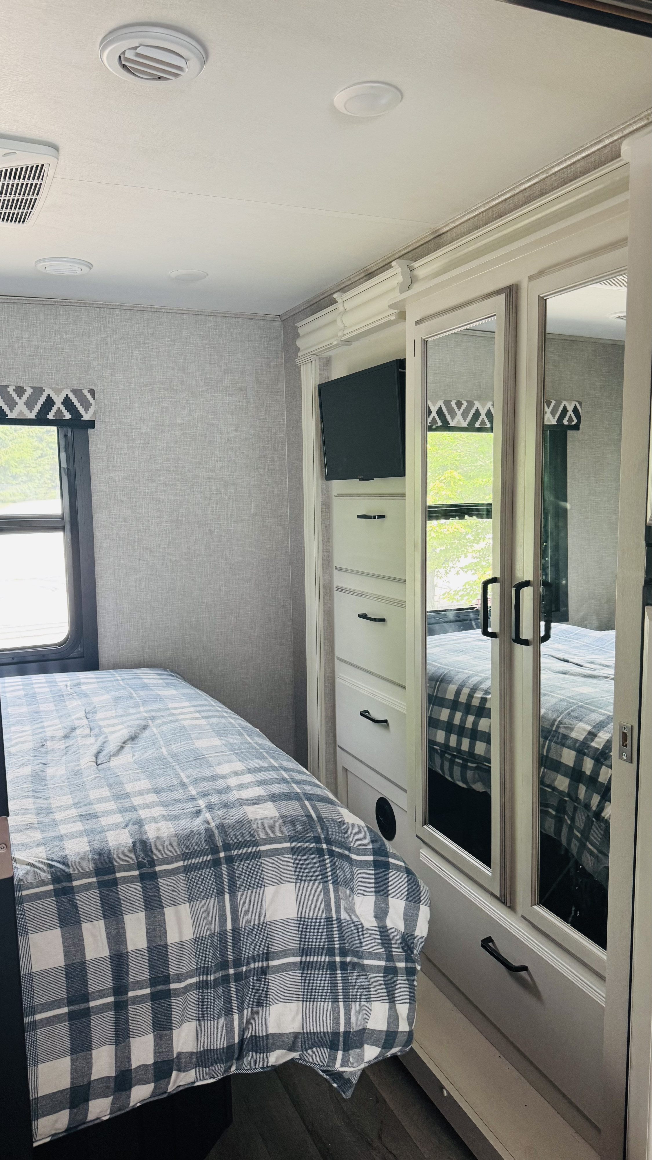 Inside an RV rental bedroom: cozy plaid bed, mirrored cabinet, TV, and wall-mounted air vent for comfortable travel.. Jayco Alante 2022