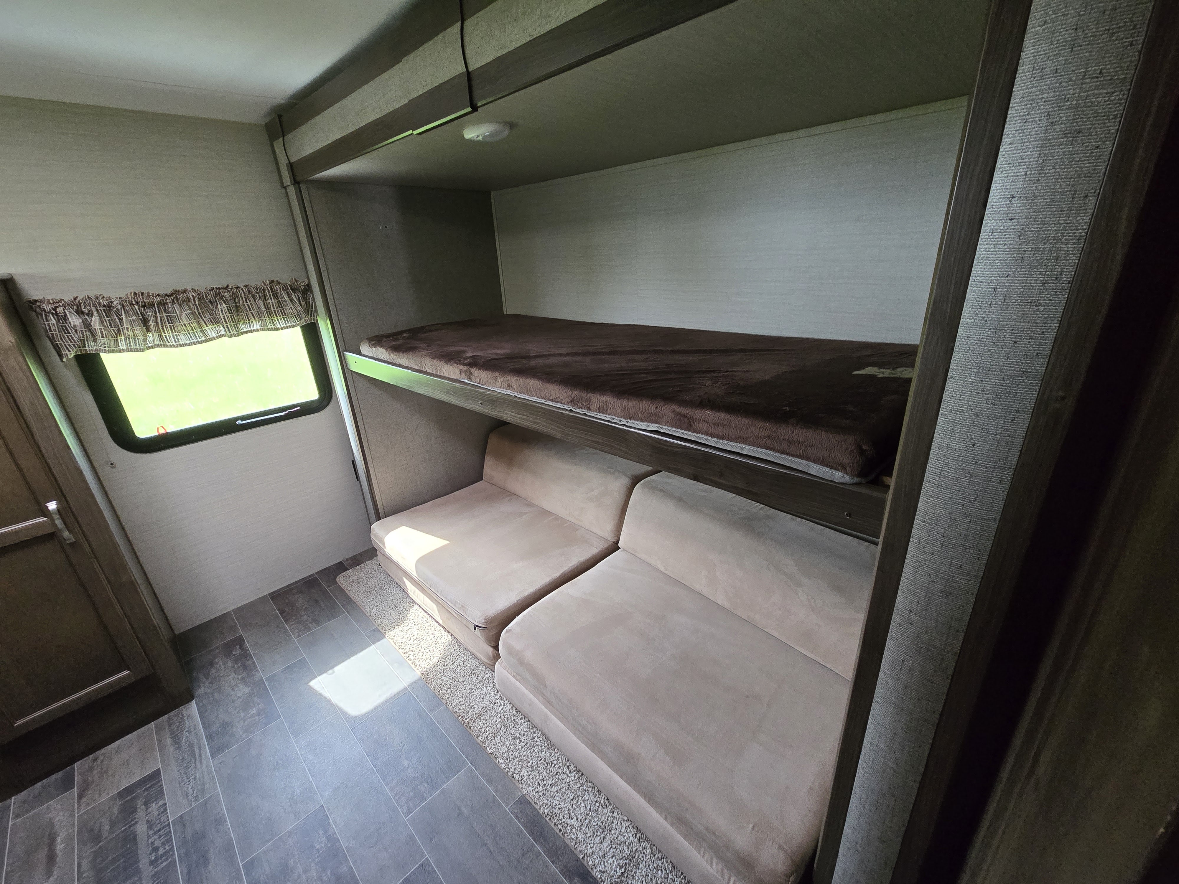 An RV rental interior showing a top bunk bed with mattress, a lower modular sofa, window, and tiled floor.. Keystone RV Bullet Ultra Lite 2018
