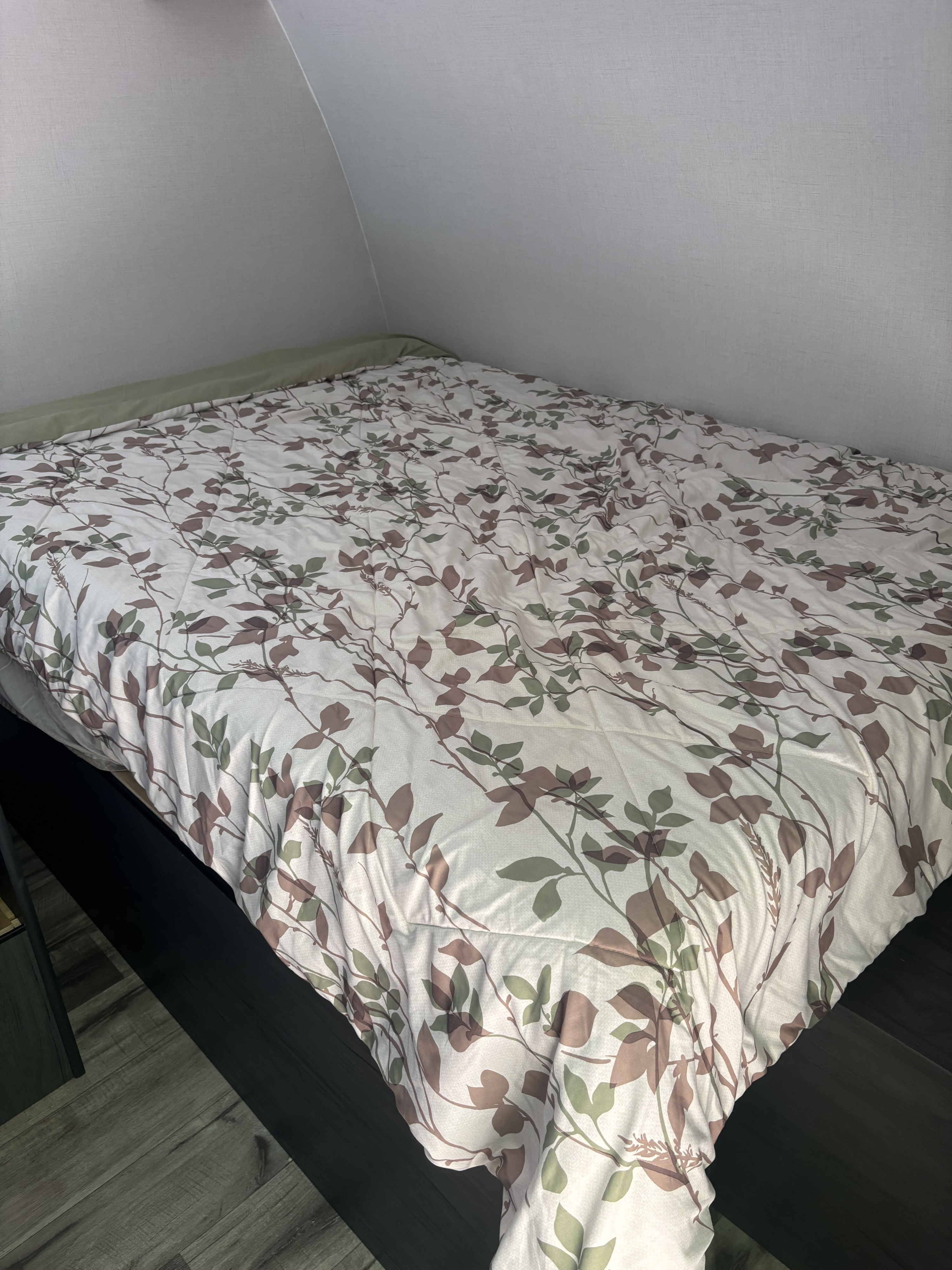 Inside an RV rental, a comfy bed with a white and green leaf patterned comforter. The curved ceiling shows it's a compact travel space.. Dutchman Aspen Trail 2025