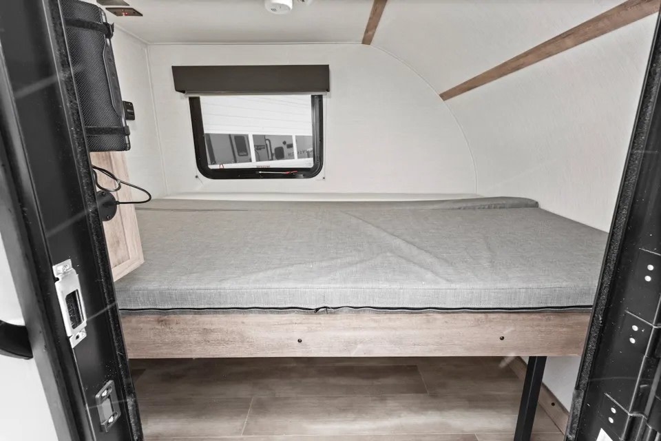 Inside this cozy RV rental: a compact bed with gray mattress, window, and curved white walls with wood accents.. GulfStream Trail Boss 1090K 2025