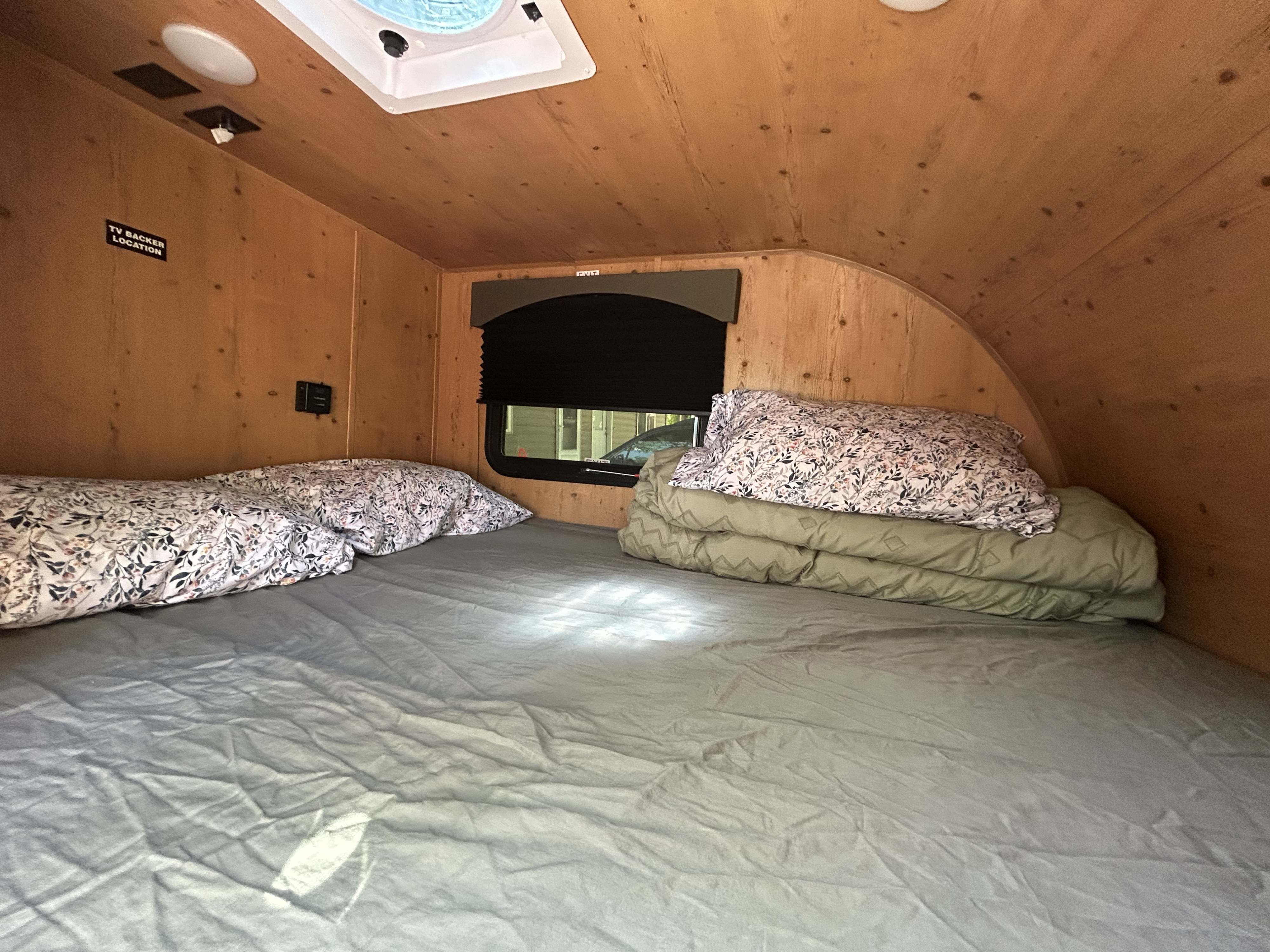 Cozy RV rental interior: wood-paneled sleeping area with bed, floral pillows, ceiling vent, and TV backer location.. Sunset Park RV SunRay 2024