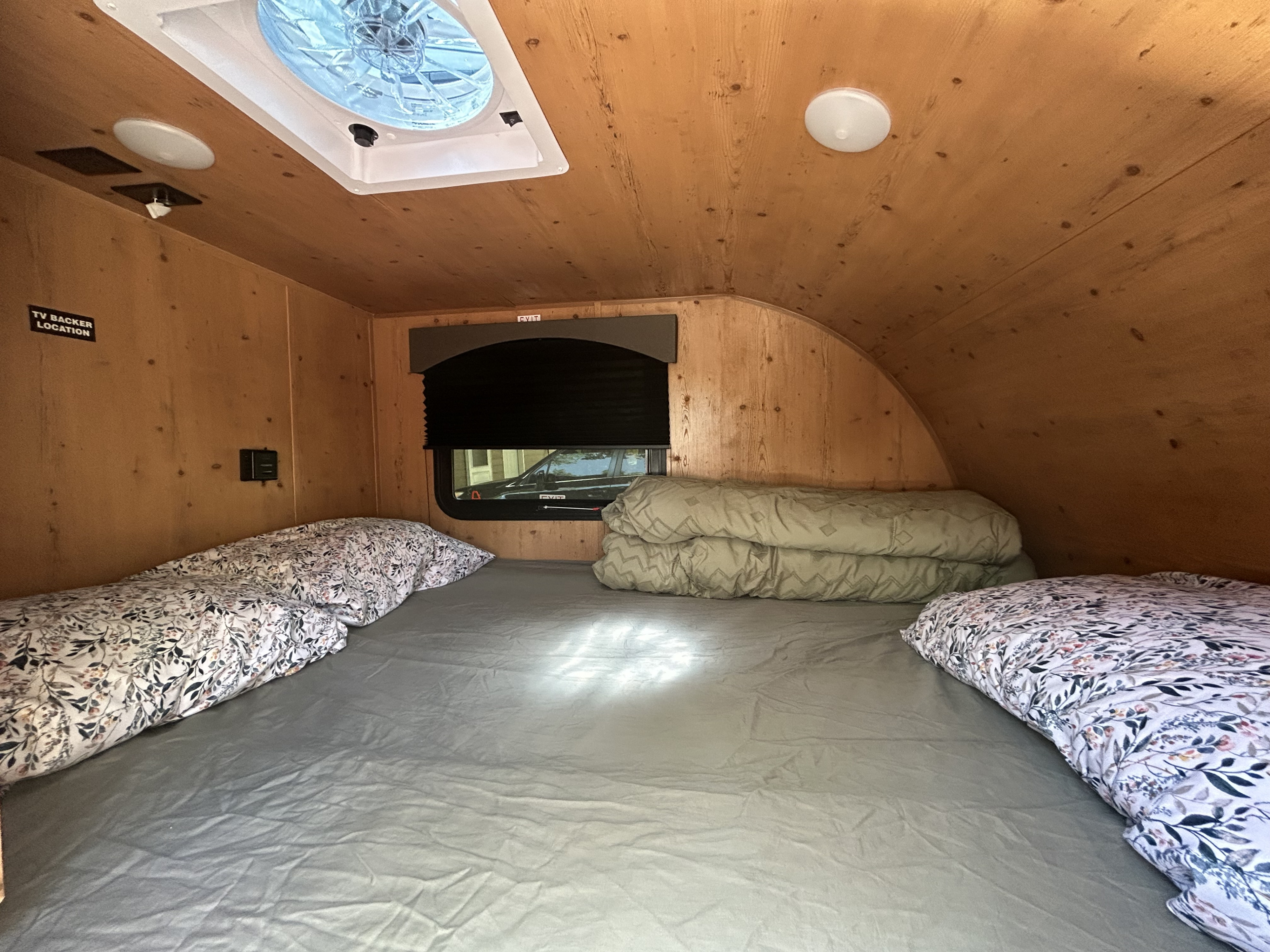 Comfortable bed and wood-paneled interior of an RV rental, showing the sleeping area with a fan and window.. Sunset Park RV SunRay 2024