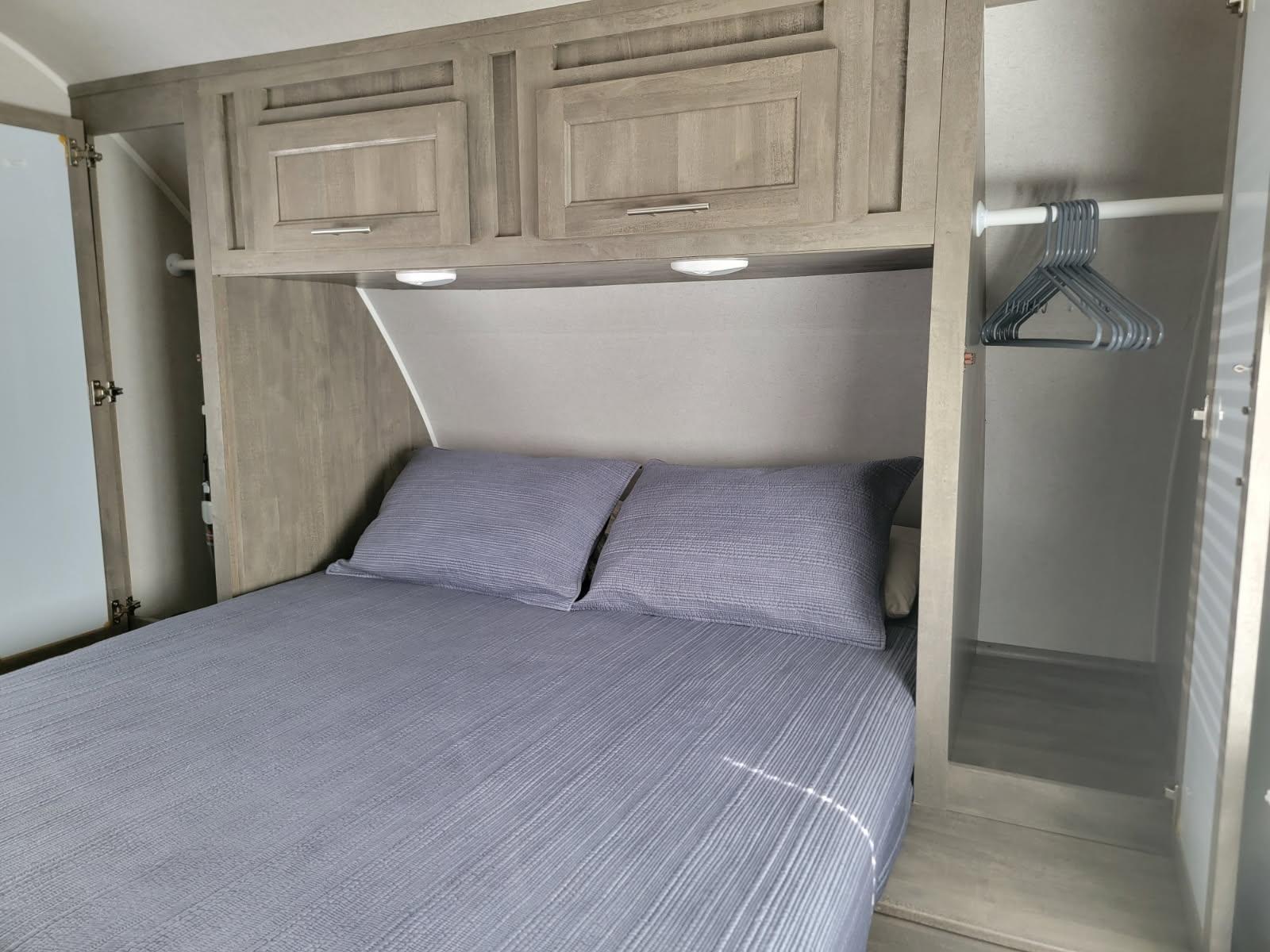 Inside an RV rental: a cozy bedroom with blue bedding, overhead storage, reading lights, and side hanging closets.. Forest River Vibe 2020