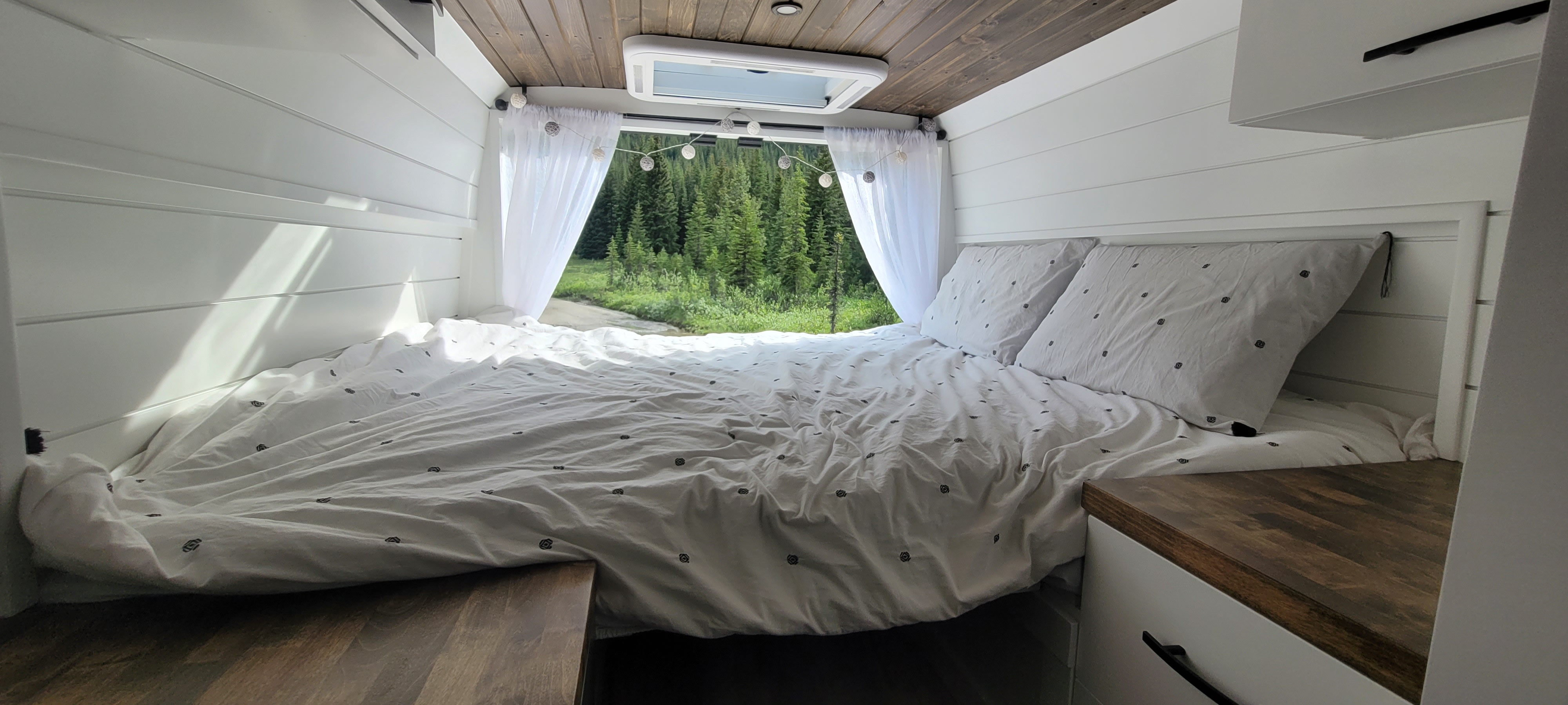 Spacious RV rental interior features a large bed with white patterned bedding. Rear window offers a lush forest and road view.. Ram Promaster 3500 Extended 2025