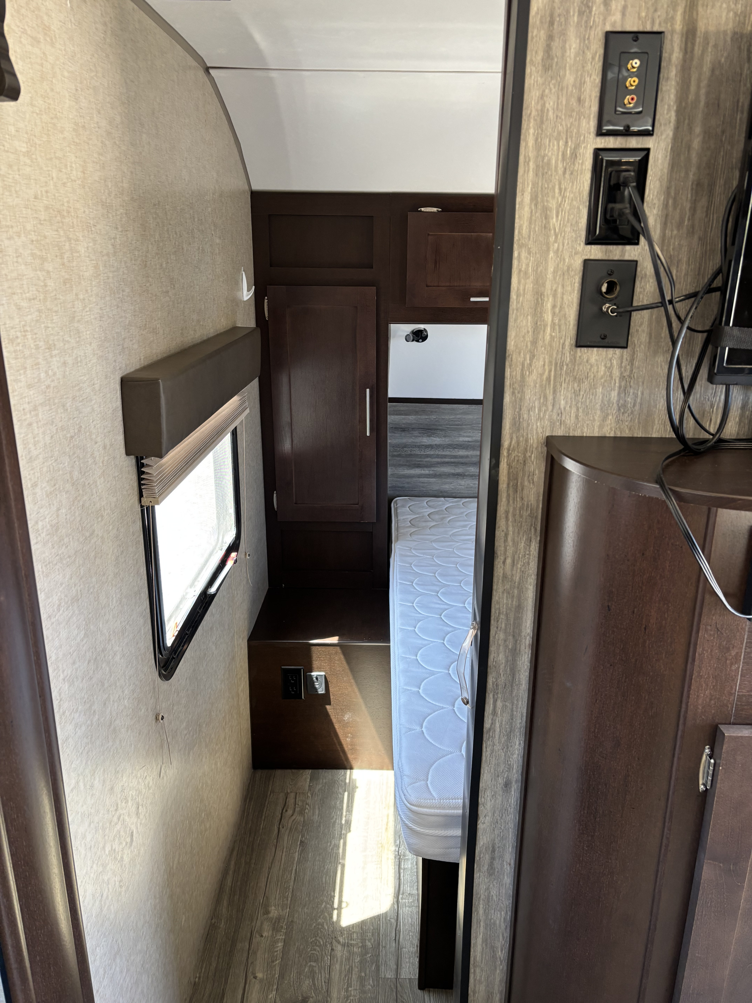 Inside this RV rental: A narrow hallway leads to a bedroom with a white mattress. Dark cabinets, window, and wall outlets are visible.. Forest River Cherokee 2017