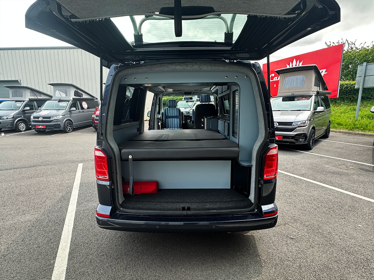 Rear view of an RV rental camper van with its hatch open, revealing a black bed and light interior. Other vans are visible.. Volkswagen T6 LWB CamperKing Portofino 2018