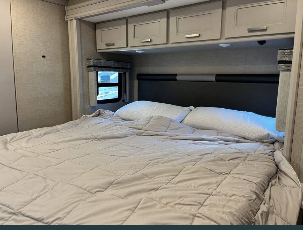 Cozy RV rental bedroom featuring a comfy bed with light grey bedding, overhead cabinets, and a small side window.. Thor Miramar 2023