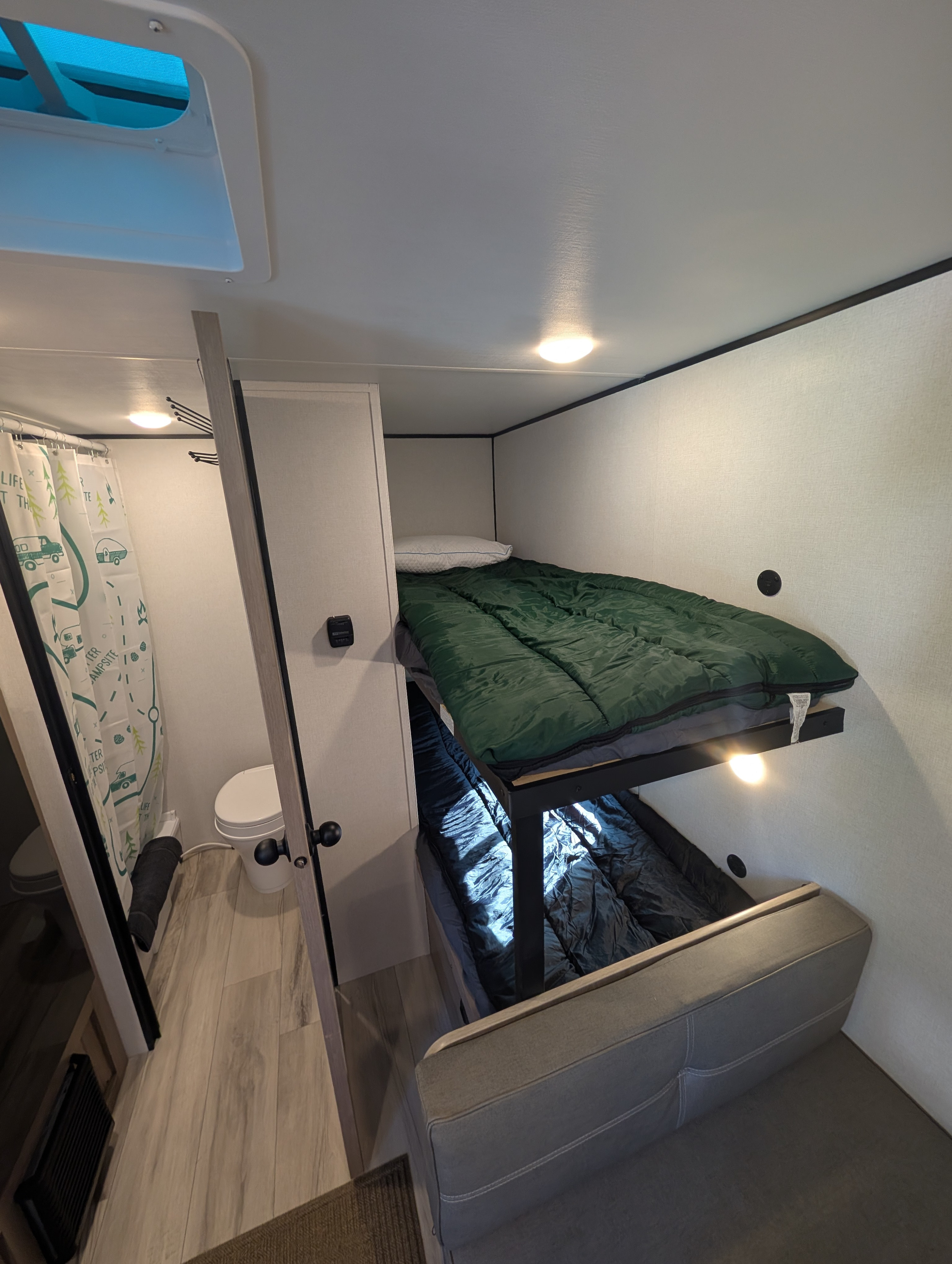 RV rental interior view: stacked bunk beds with sleeping bags, a compact bathroom to the left, and a gray sofa.. Coleman 17B 2025
