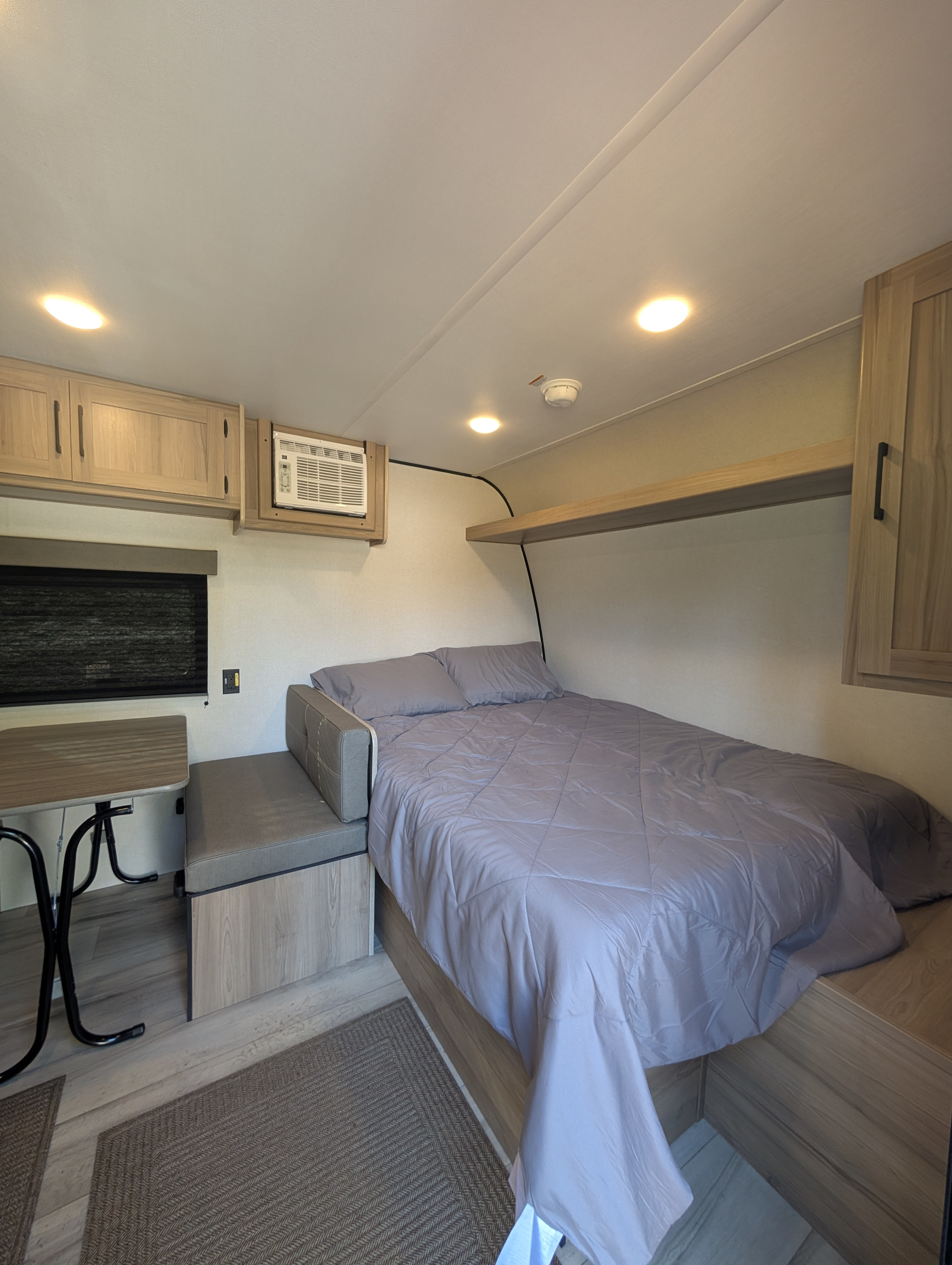 Inside a modern RV rental: a neat bed, compact dinette, AC unit, and ample overhead storage. Ready for travel!. Coleman 17B 2025