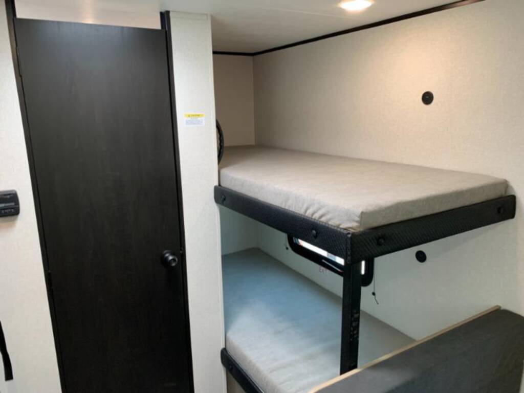 Inside an RV rental: light walls frame sturdy bunk beds with light mattresses, beside a dark, textured privacy door.. Jayco Jay Flight 2026