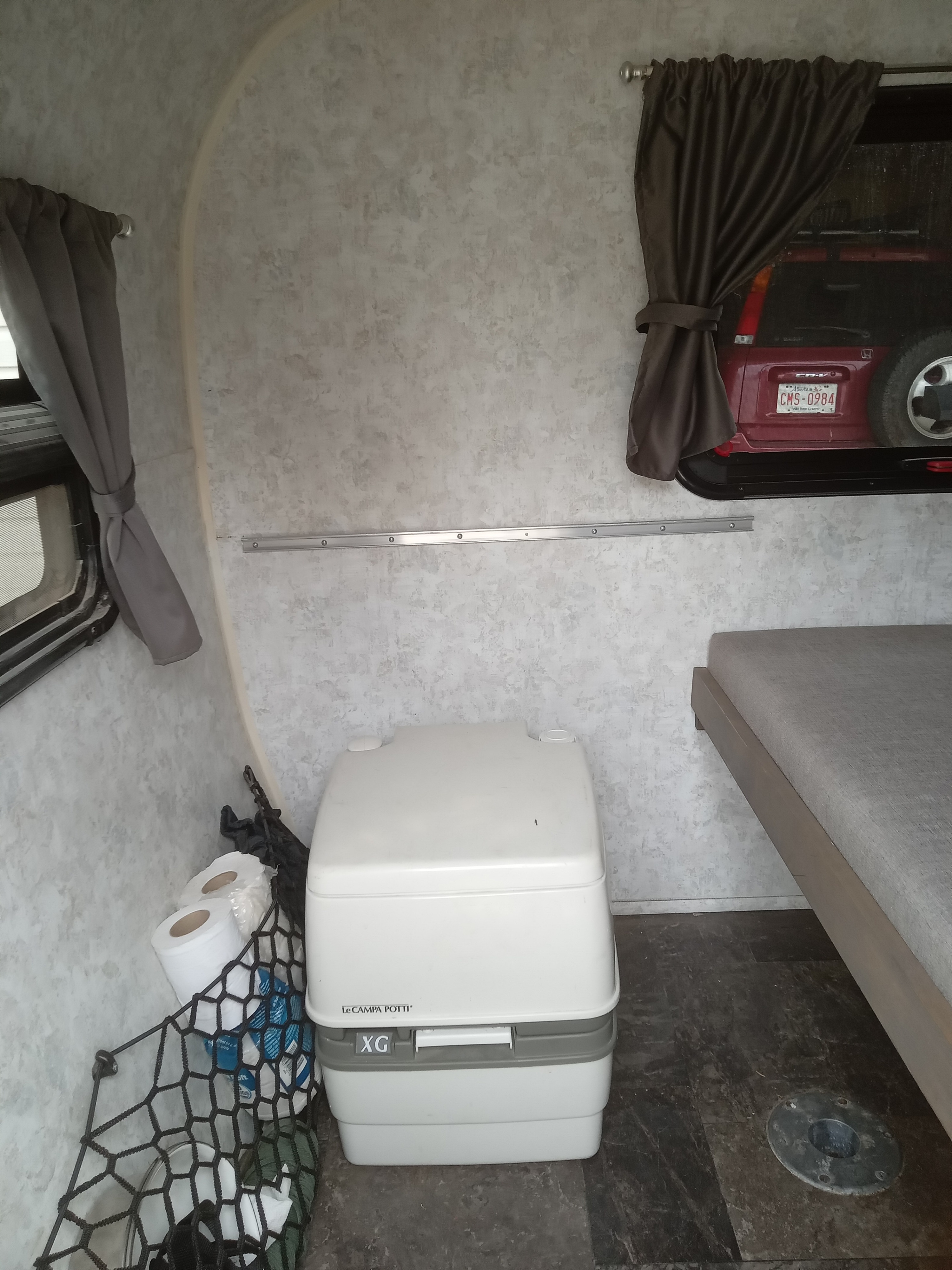 RV rental interior: portable toilet, bed, windows, a storage net with supplies, and a red car visible outside.. Prolite Prolite Trailer 2018