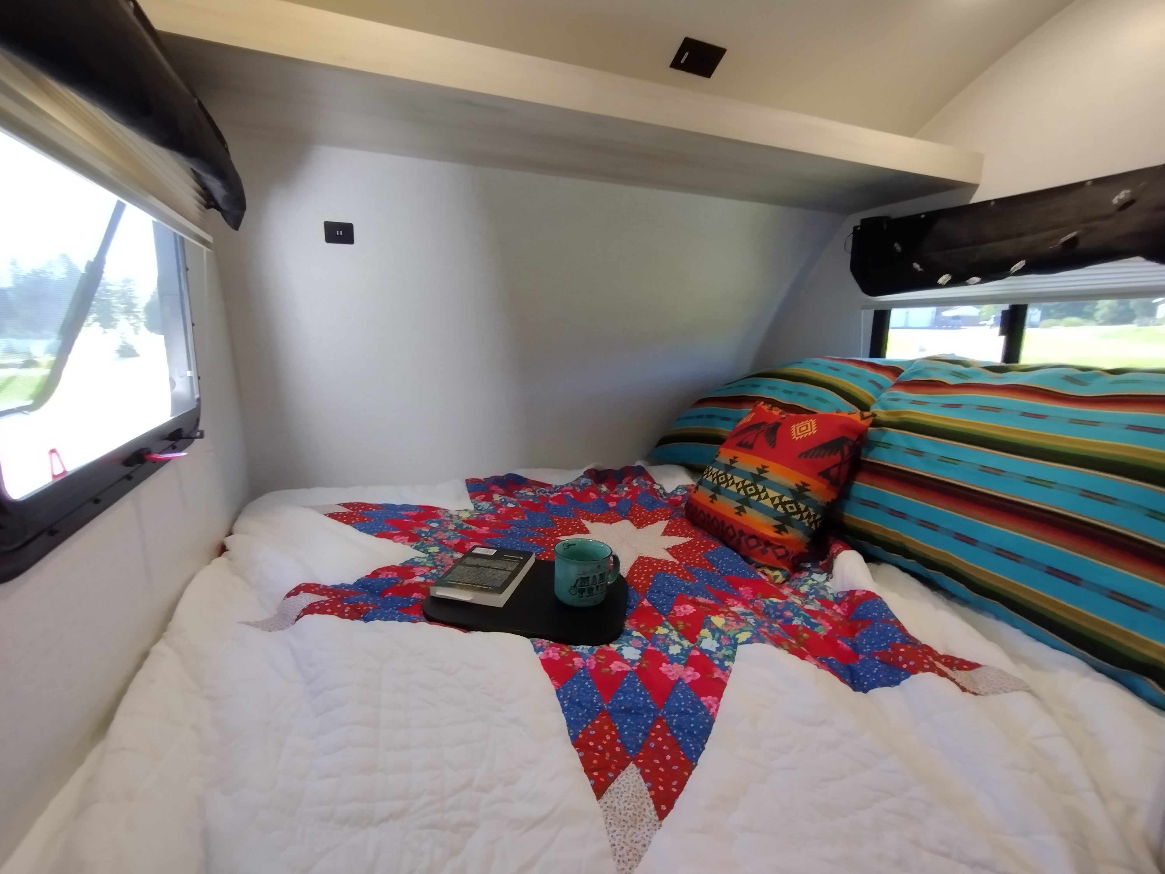 Cozy RV rental bedroom featuring a colorful quilt, pillows, and a reading nook with a book and mug.. Winnebago Hike 2022