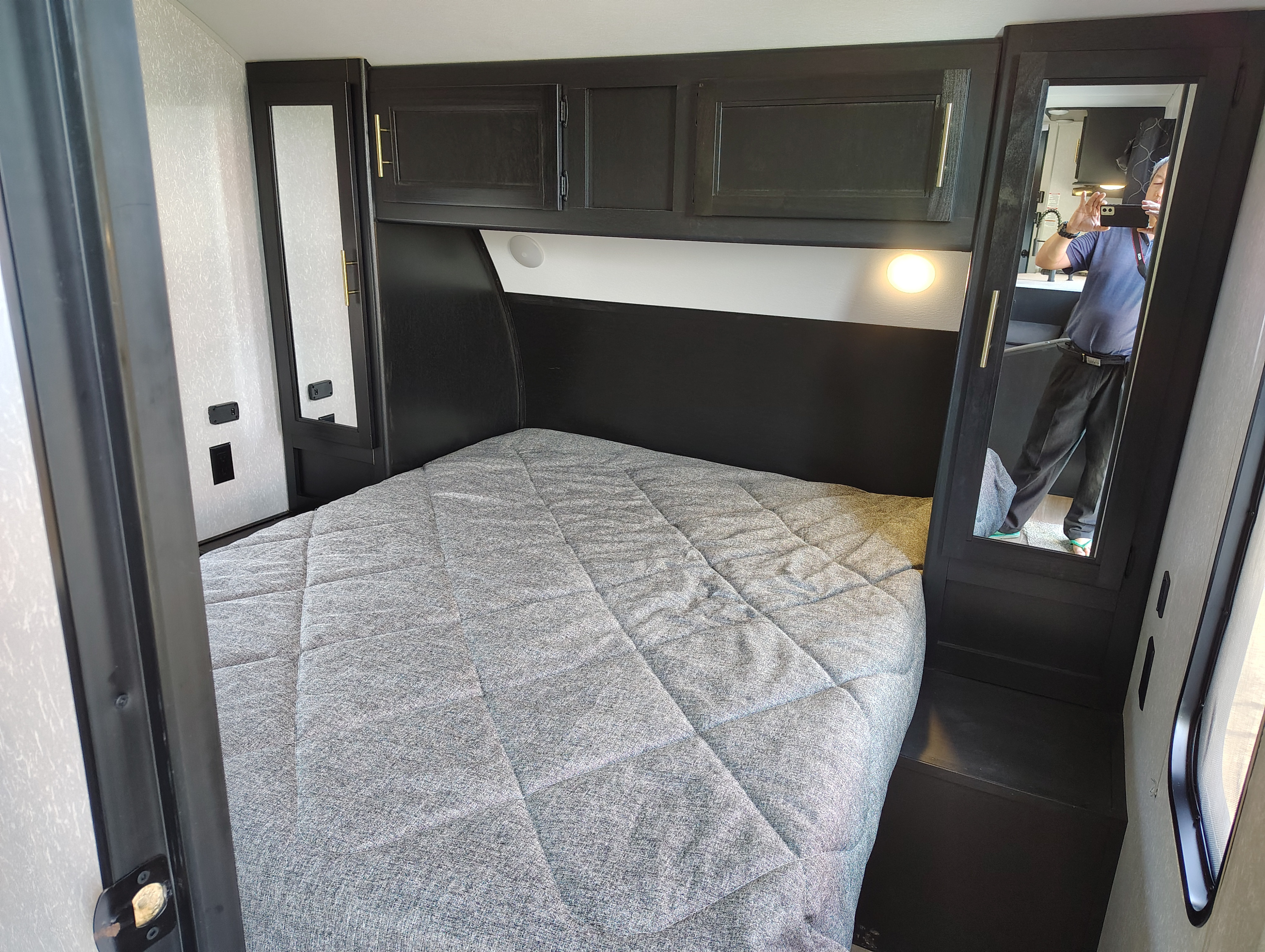 Inside an RV rental: a compact bedroom with dark cabinets, gray quilted bed, built-in light, and mirror.. Forest River Cherokee 2022