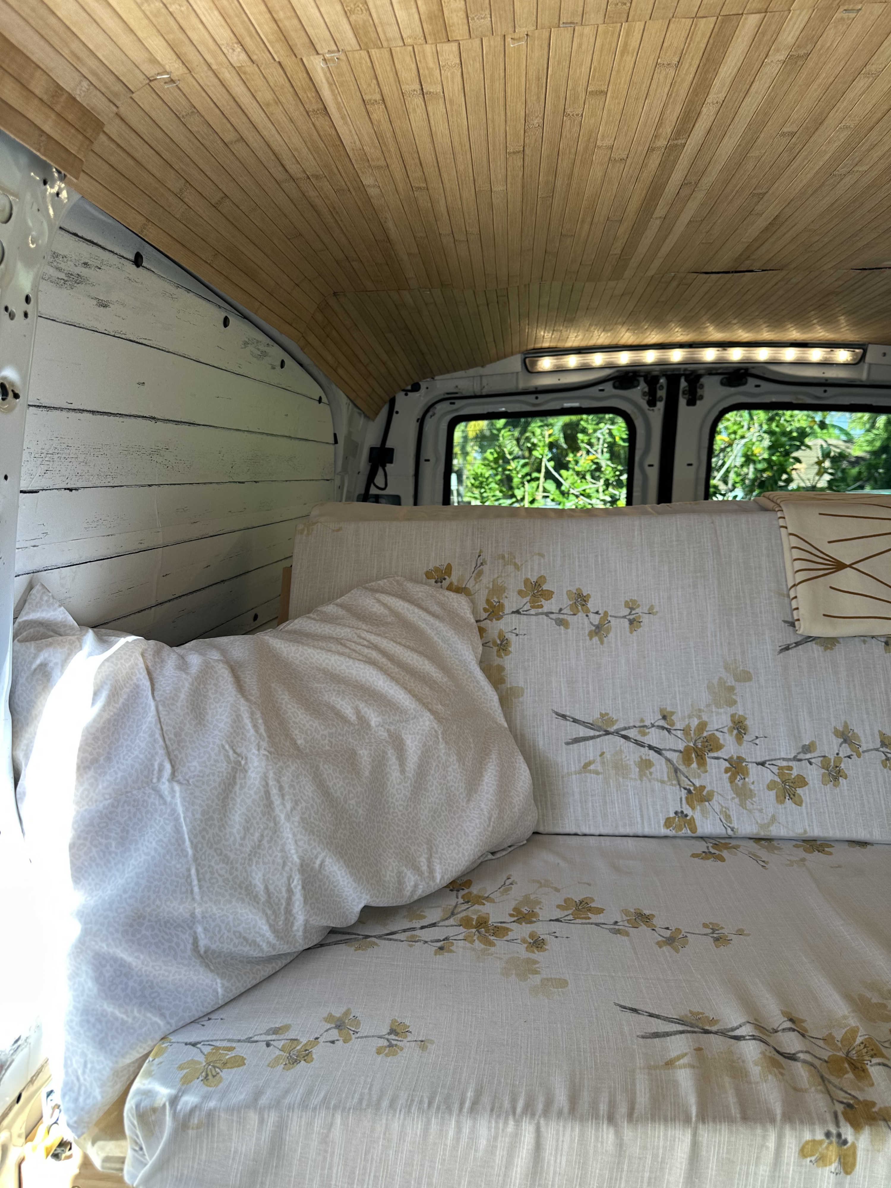 Cozy RV rental interior featuring a wood-slat ceiling, white paneling, floral bed, and bright rear window view.. Mercedes-Benz Metris 2020