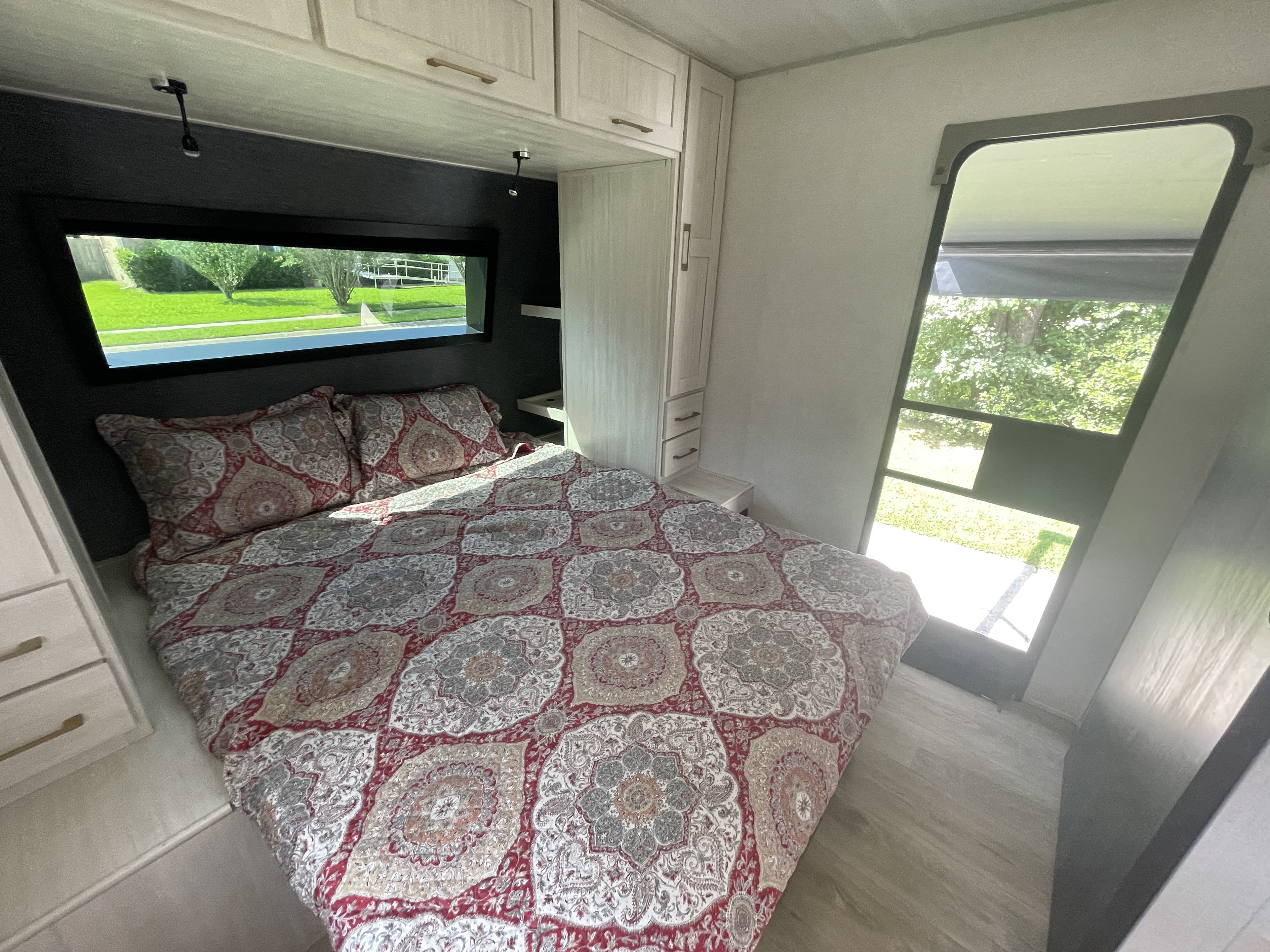 Cozy RV rental bedroom featuring patterned bedding, overhead cabinets, and a scenic window view of green outdoor space.. Forest River Grand Surveyor 2024