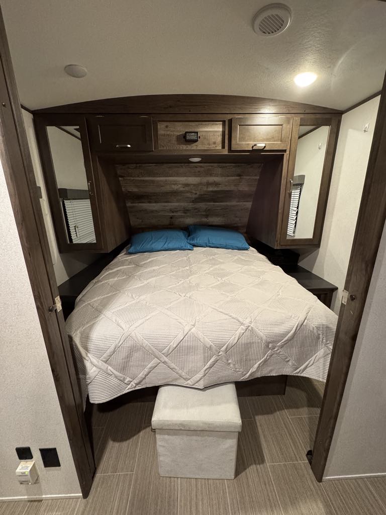 Cozy RV rental bedroom features a comfy bed, wood headboard, overhead storage, and side mirrors, optimizing space.. Keystone RV 3400QD 2020