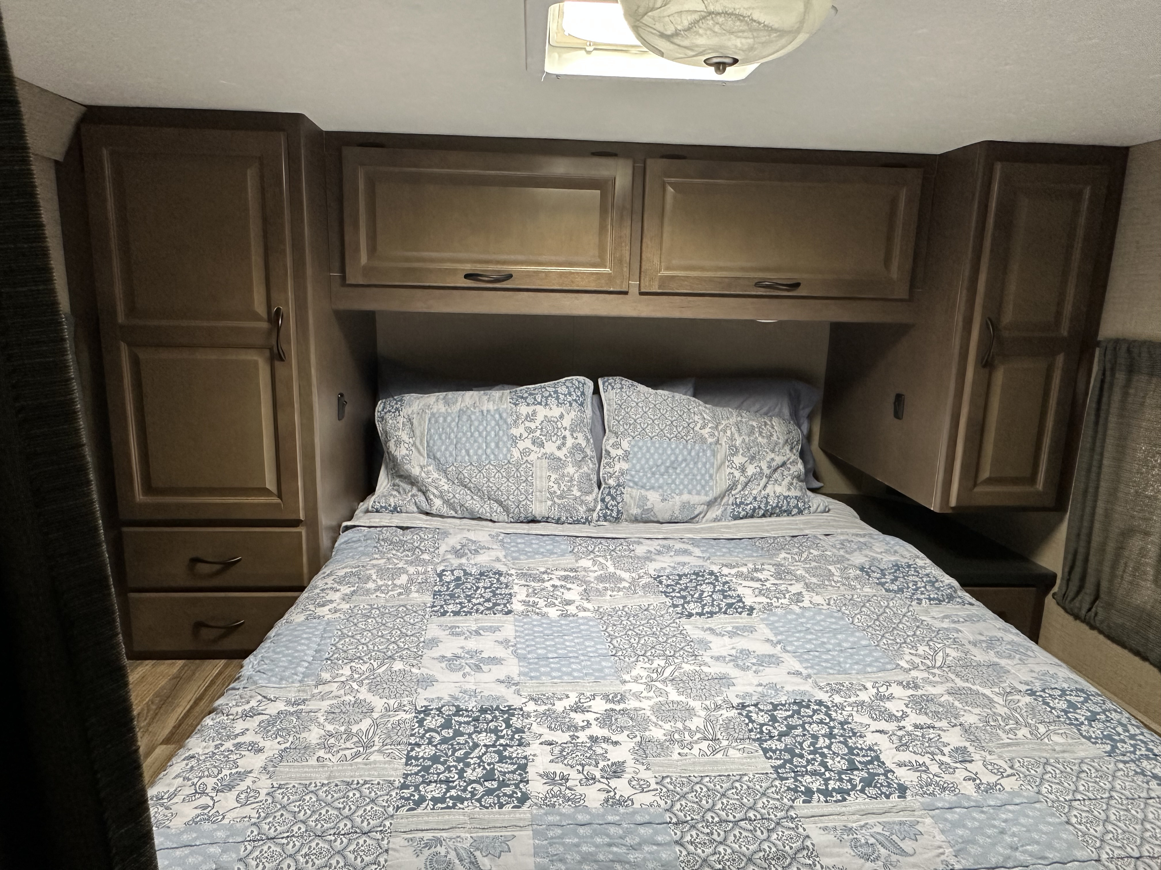 Inside an RV rental bedroom: a cozy bed with blue patchwork bedding, surrounded by wooden overhead and side storage cabinets.. Thor Majestic 2019