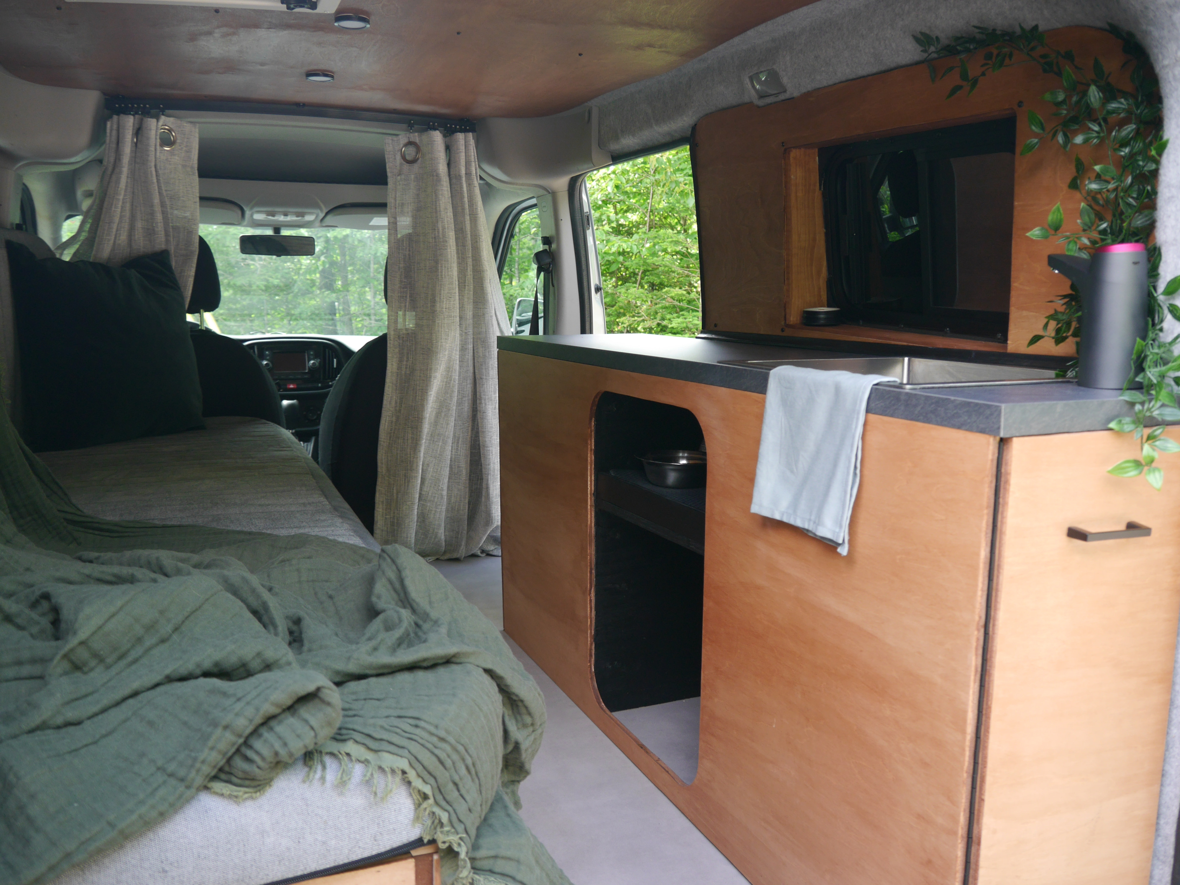 RV rental interior: a converted camper van with a cozy bed, compact kitchen unit, and lush green views.. Ram Promaster City 2015