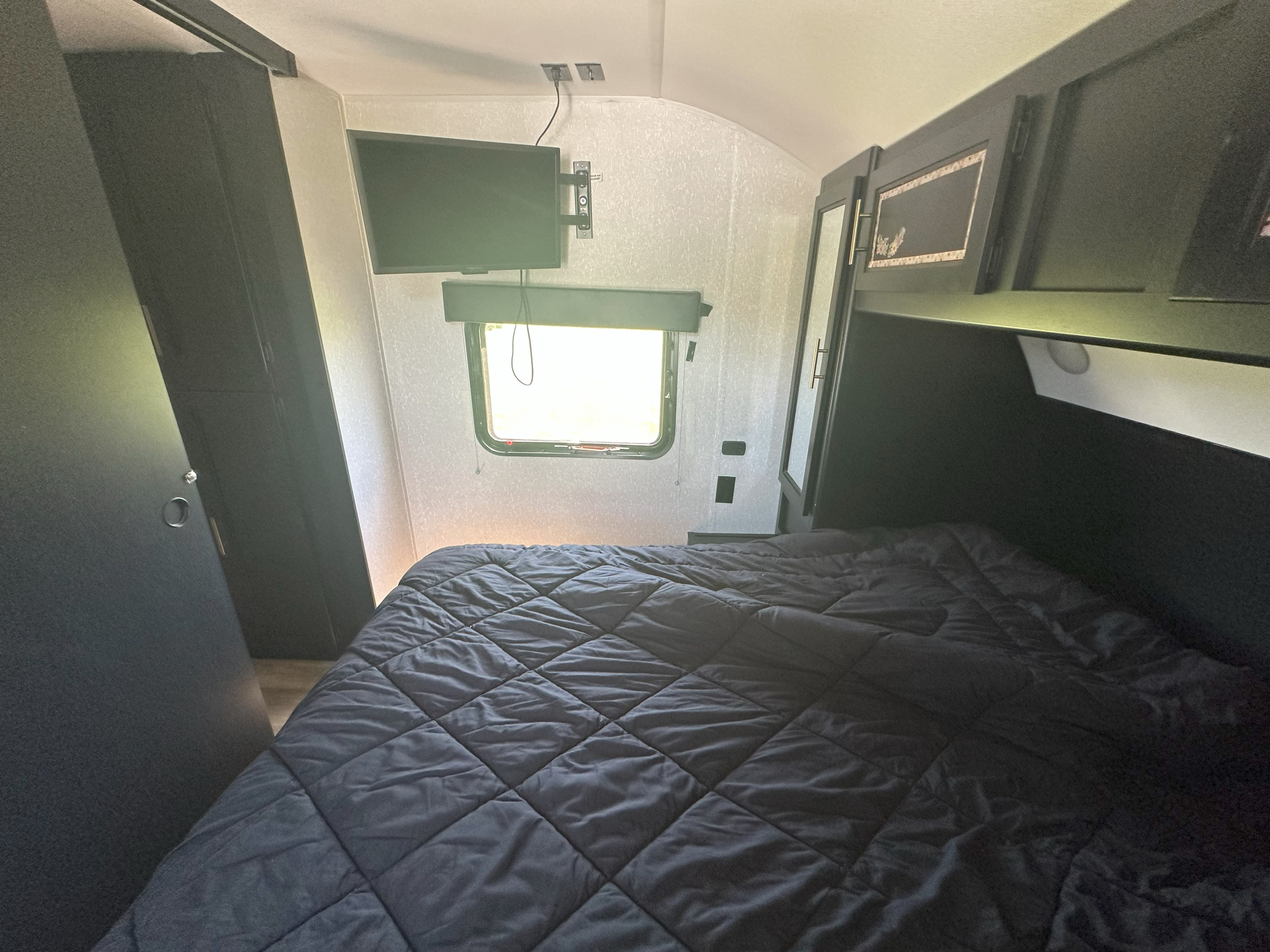 Cozy RV rental bedroom features a dark comforter bed, wall-mounted TV, window, and ample storage cabinets.. Forest River Cherokee 2022