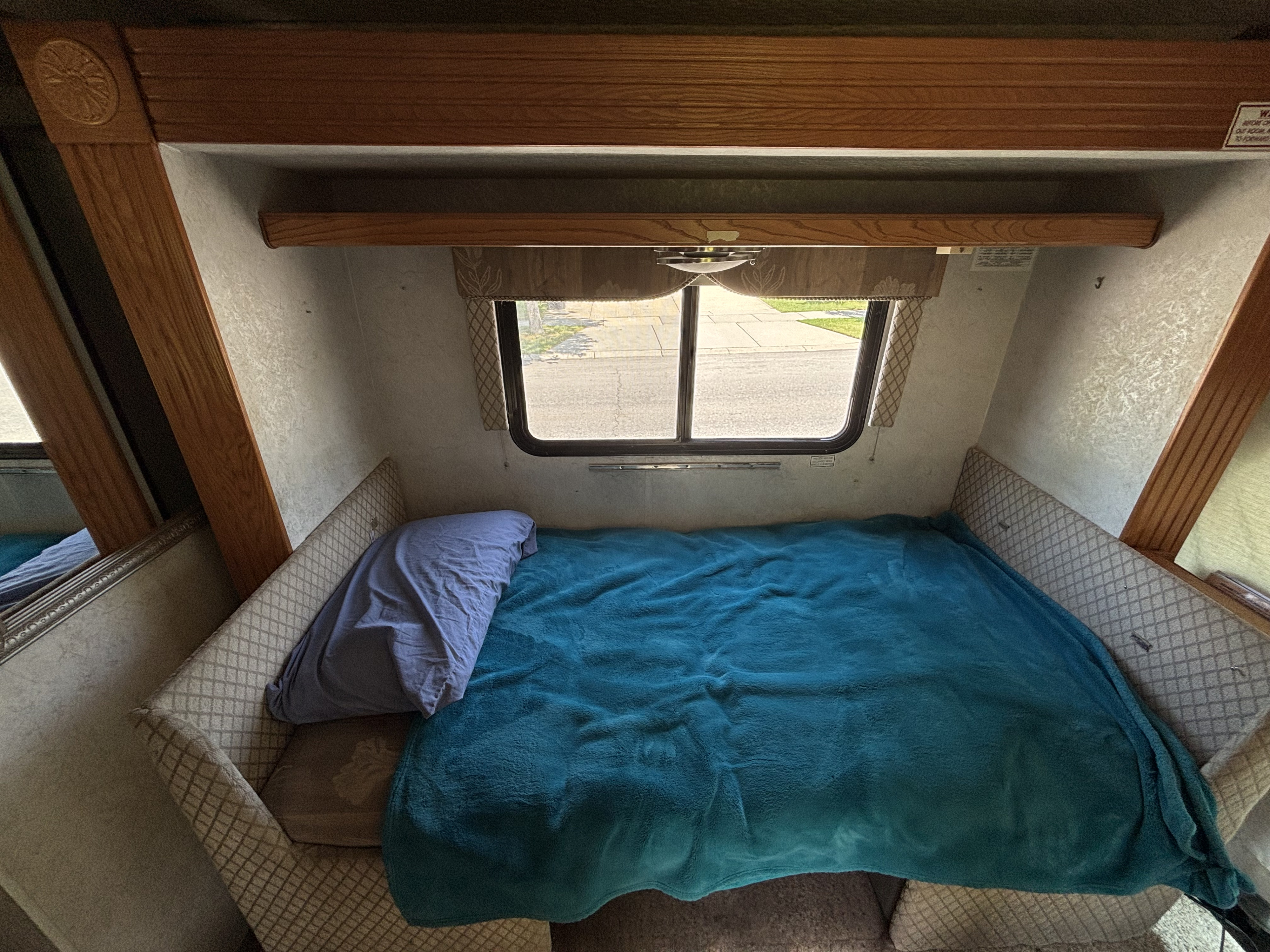 Cozy RV rental interior shows a sleeping area with a teal blanket, purple pillow, and window view.. Gulf Stream BT Cruiser 2004