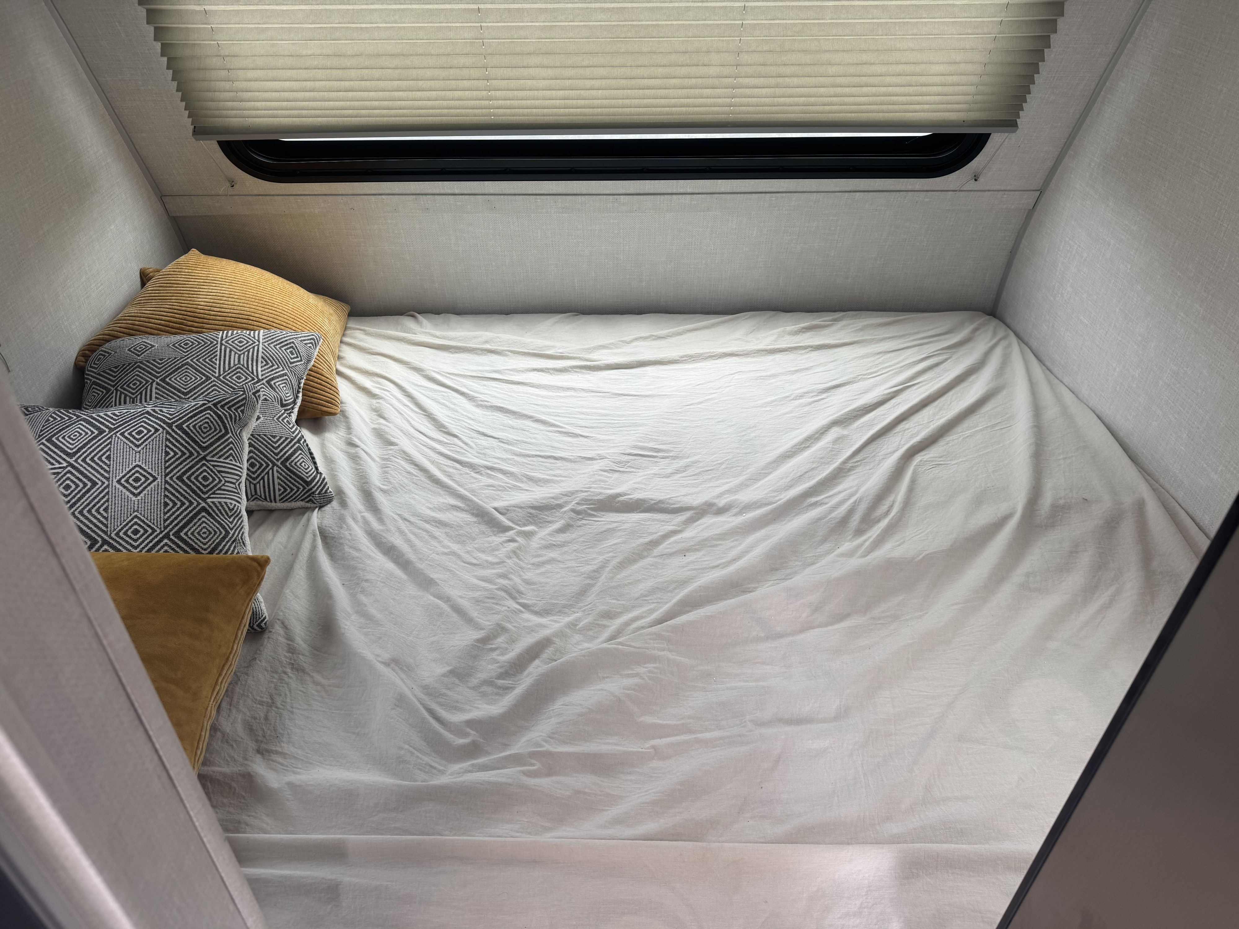 Cozy RV rental bed with white sheets, patterned and yellow pillows, and a window with pleated blinds.. No Boundaries 16 2023
