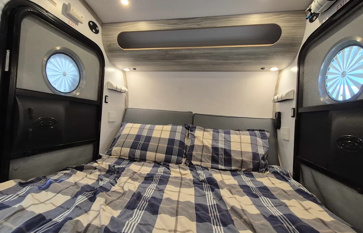 Inviting RV rental sleeping area: a bed with blue/beige plaid bedding, flanked by doors with round windows & overhead storage.. Bean Trailer Mean Bean 2022