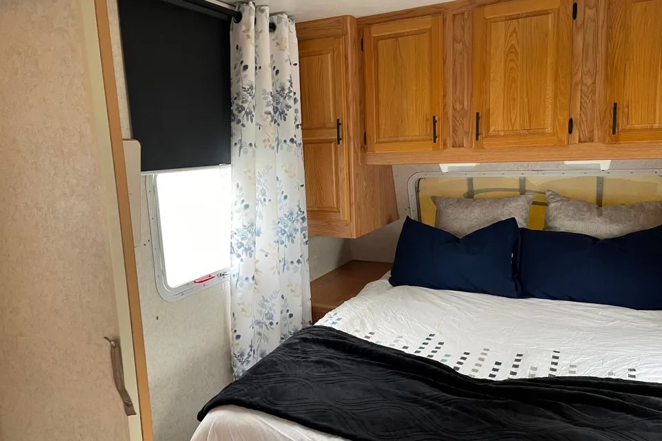 RV rental bedroom featuring a comfy bed, wood cabinets, and a window with both blackout and floral curtains.. Jayco Eagle 2002