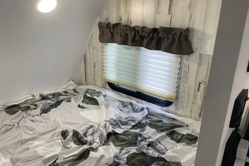 RV rental bedroom interior featuring a bed with leafy bedding, a window with a pleated blind, and a rustic wood accent wall.. Travel Lite Rove Litre 2024