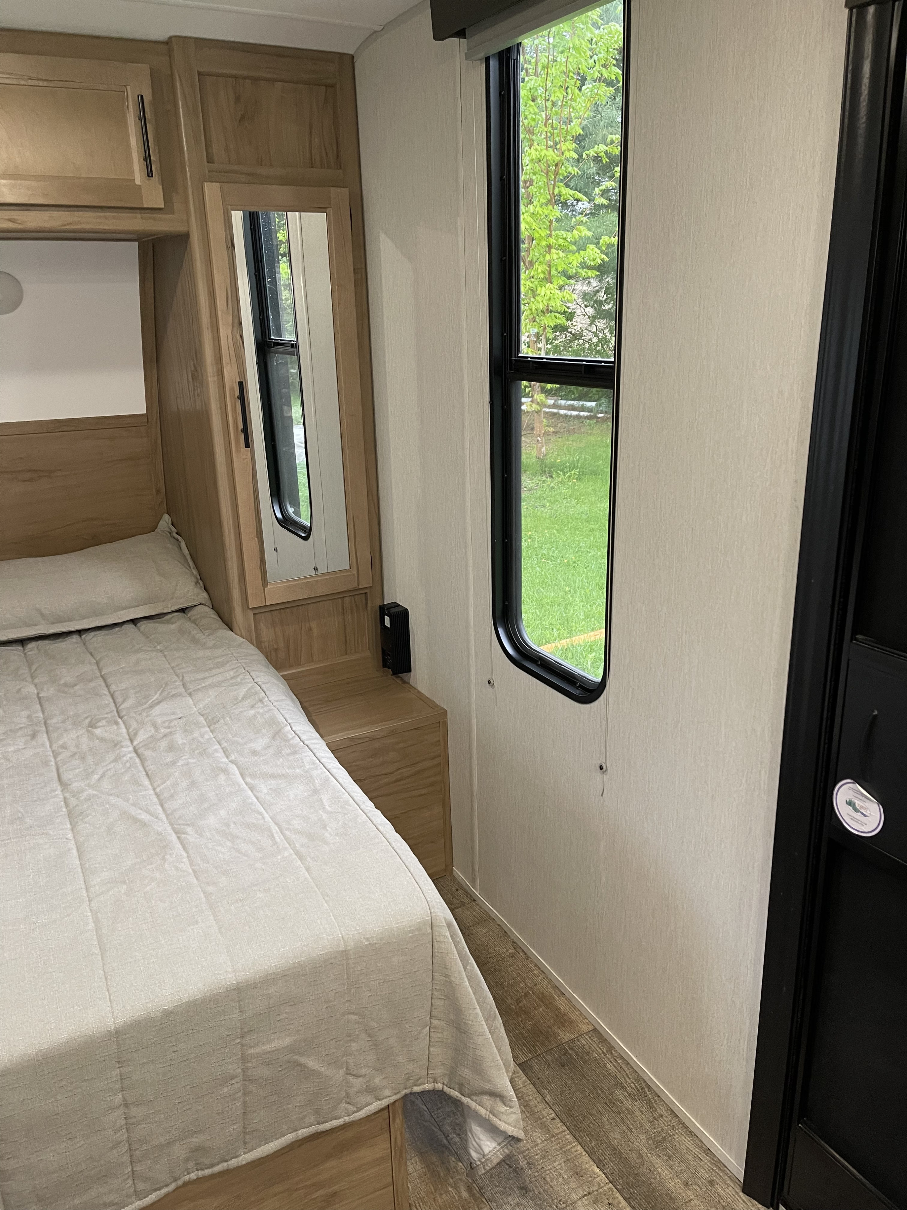 Cozy RV rental bedroom with light wood cabinetry, a comfortable bed with beige comforter, and a tall window looking out to green trees.. Forest River Cherokee Grey Wolf 2024