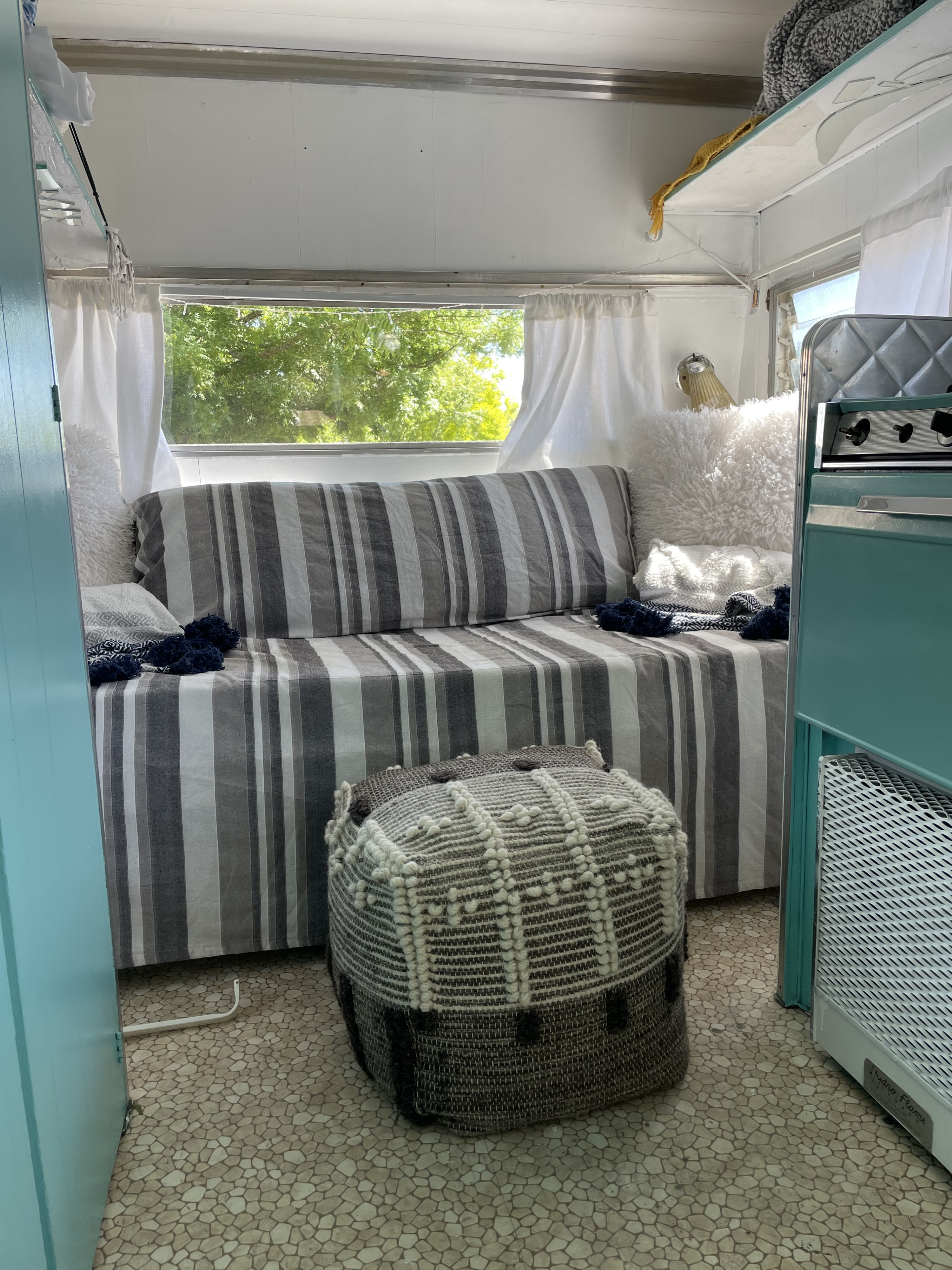 Cozy RV rental interior features a gray and white striped couch, a textured ottoman, and a bright window with green trees.. Shasta Aristocrat 1969