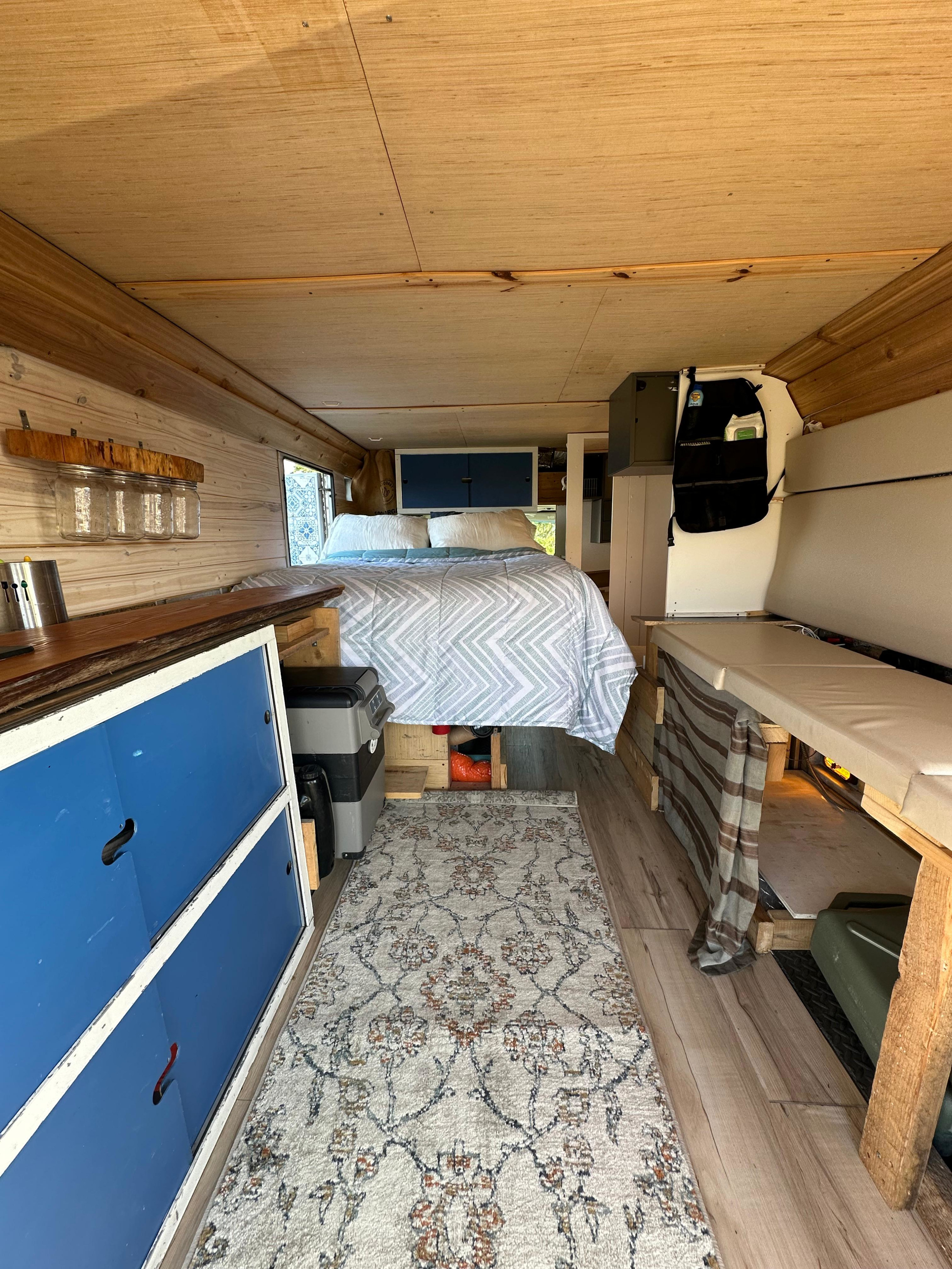Inside a cozy RV rental: wood-paneled interior with a comfy bed, kitchen counter, storage, and patterned rug.. Ford E350 Box Van 2003