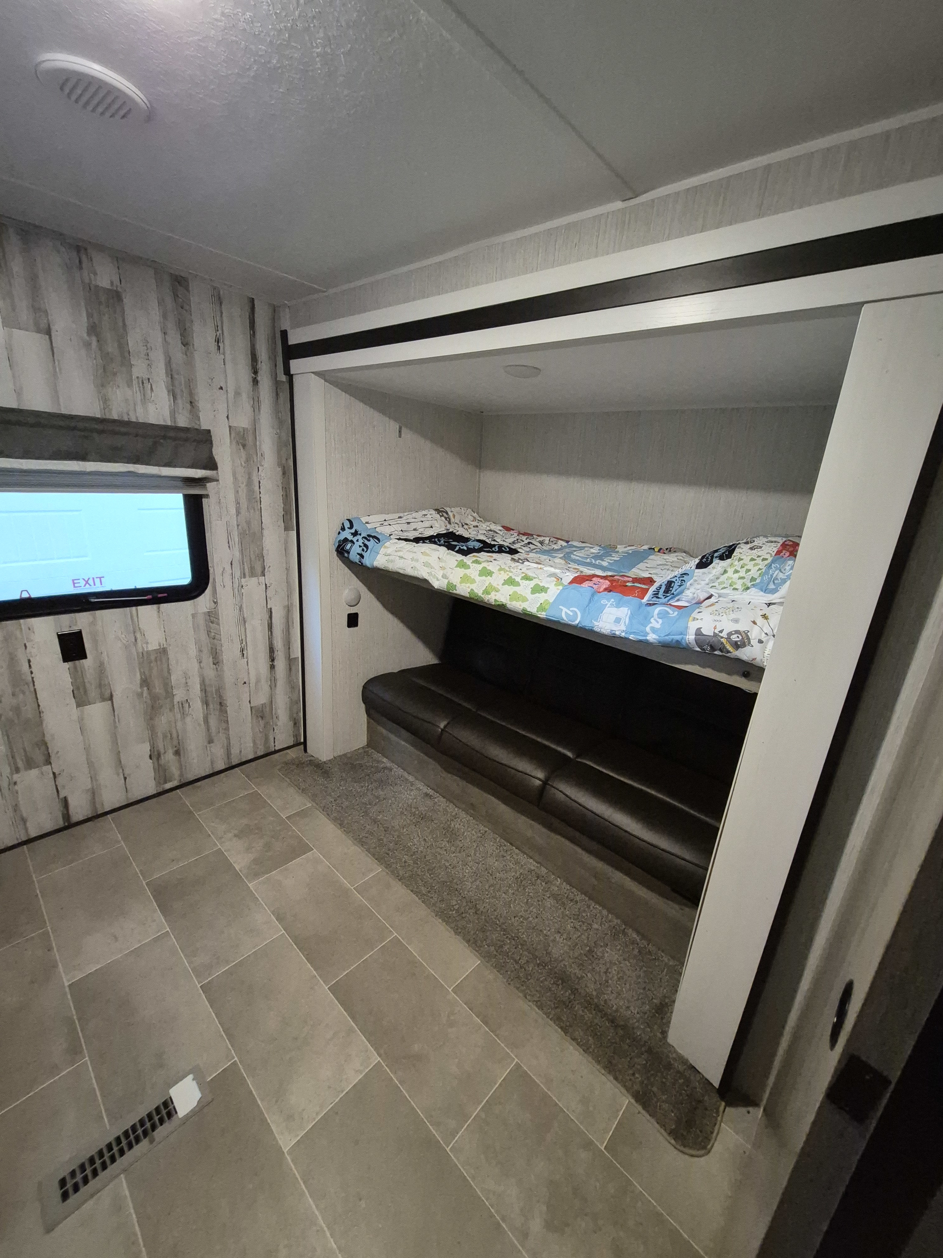 RV rental interior: cozy bunk bed with a colorful quilt above a dark sofa. Tiled floor, wood-look walls, and window with 'EXIT'.. Forest River Palomino Puma XLE Lite 2021