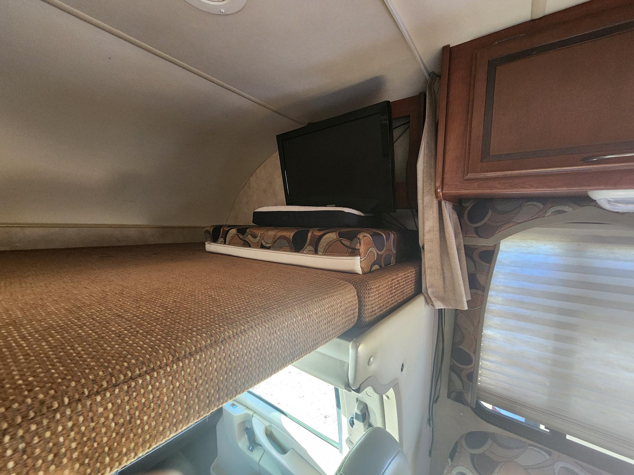 Comfortable RV rental cab-over bunk with a TV and patterned mattress, offering a cozy sleeping and lounging area inside the motorhome.. Thor Chateau 2013