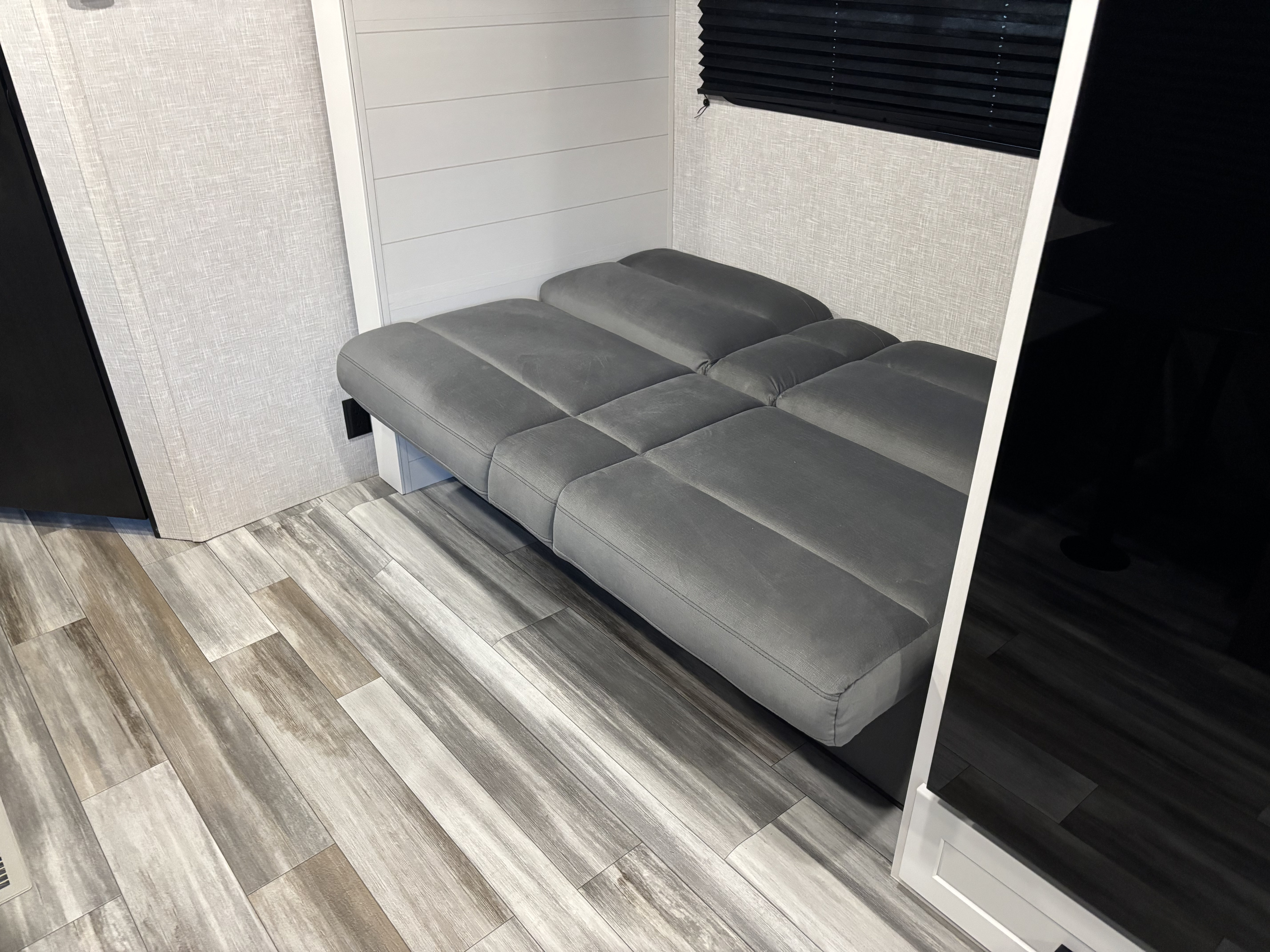 RV rental interior showcasing a multi-section gray bed/bench, light wood-look floor, textured walls, and black pleated blind.. Jayco Jay Flight SLX 2025