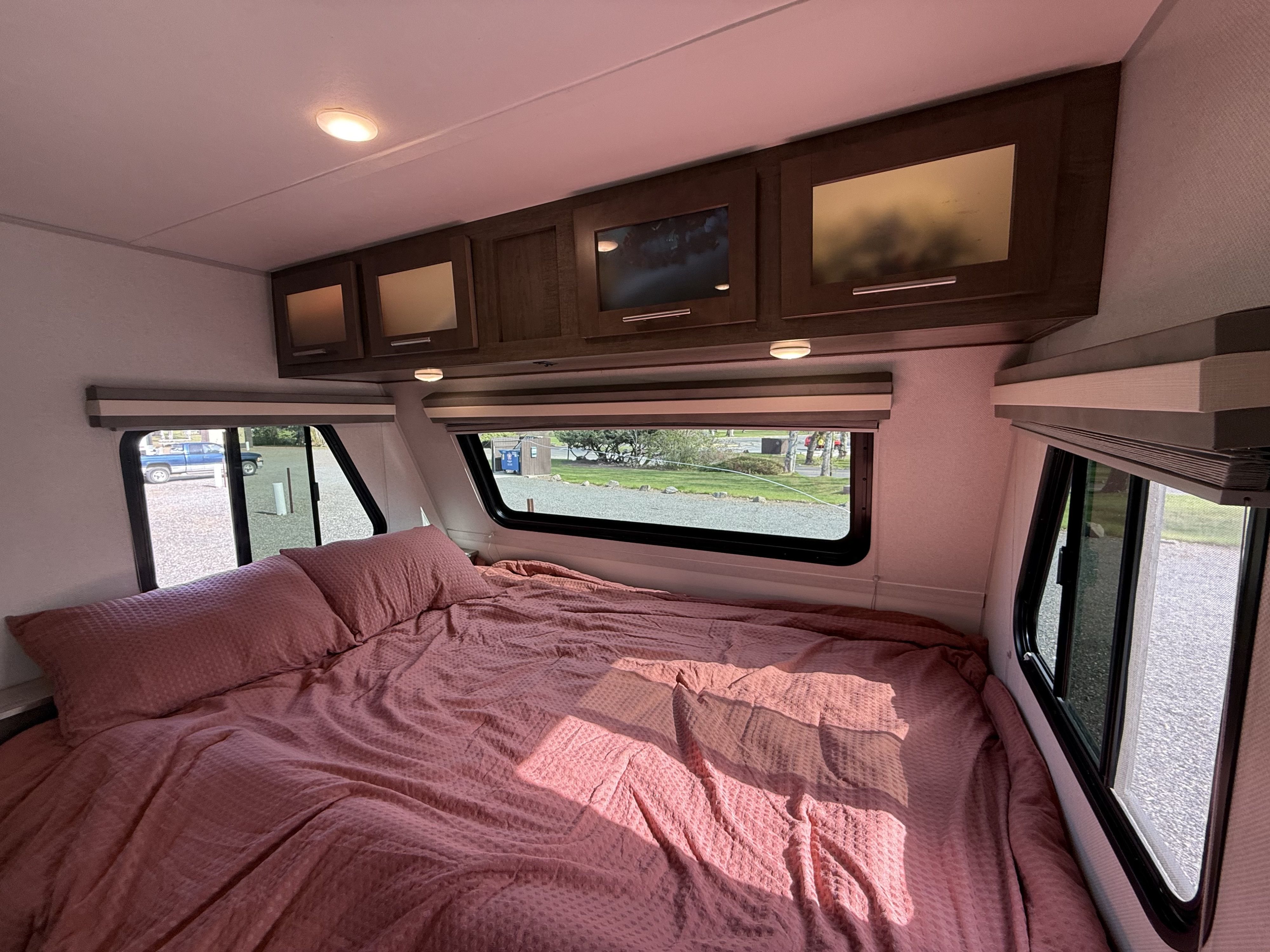 Modern RV rental bedroom featuring a comfy bed with pink sheets, overhead cabinets, and large windows revealing the campsite.. Forest River No Boundaries 2019