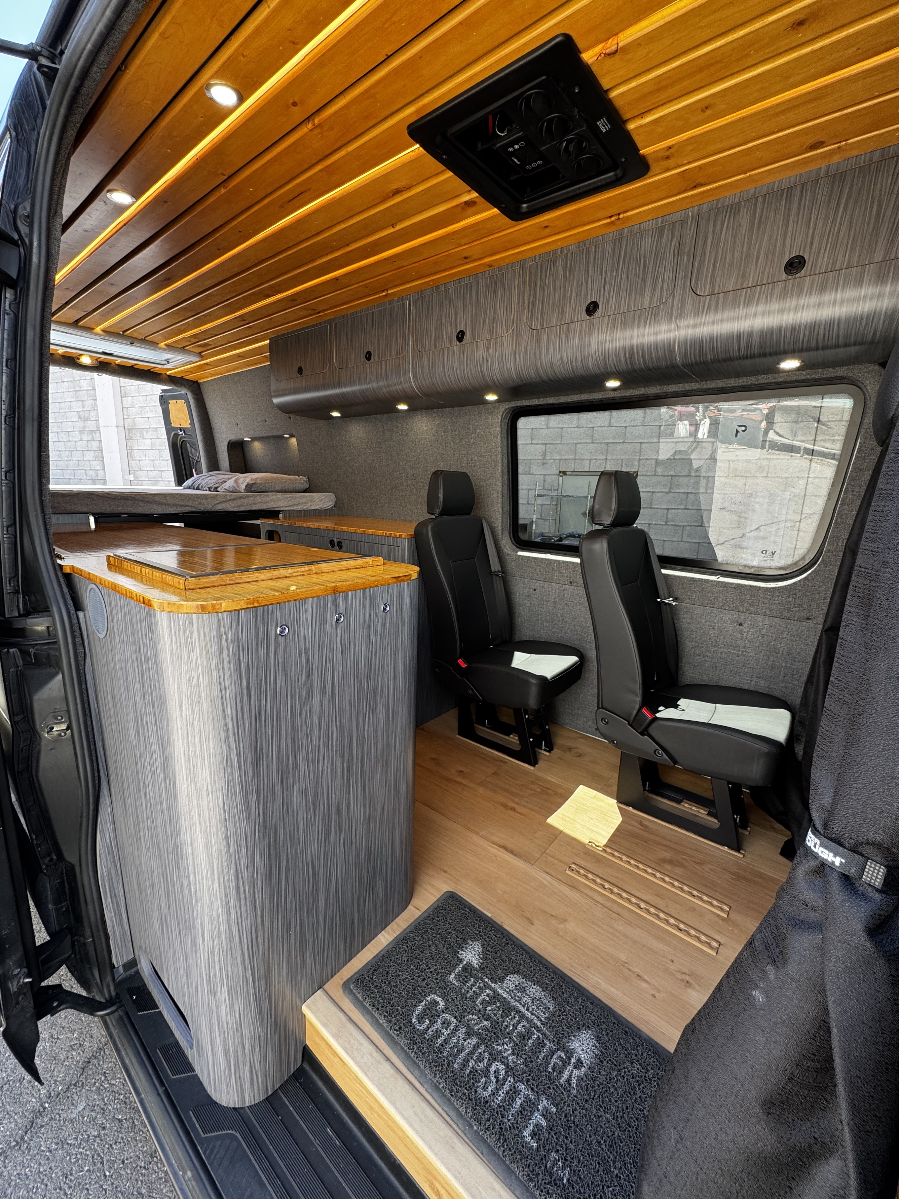 Modern RV rental interior: wood slat ceiling, gray cabinets, bed, kitchen counter, & passenger seats. Ready for adventure.. Mercedes-Benz Sprinter 2023