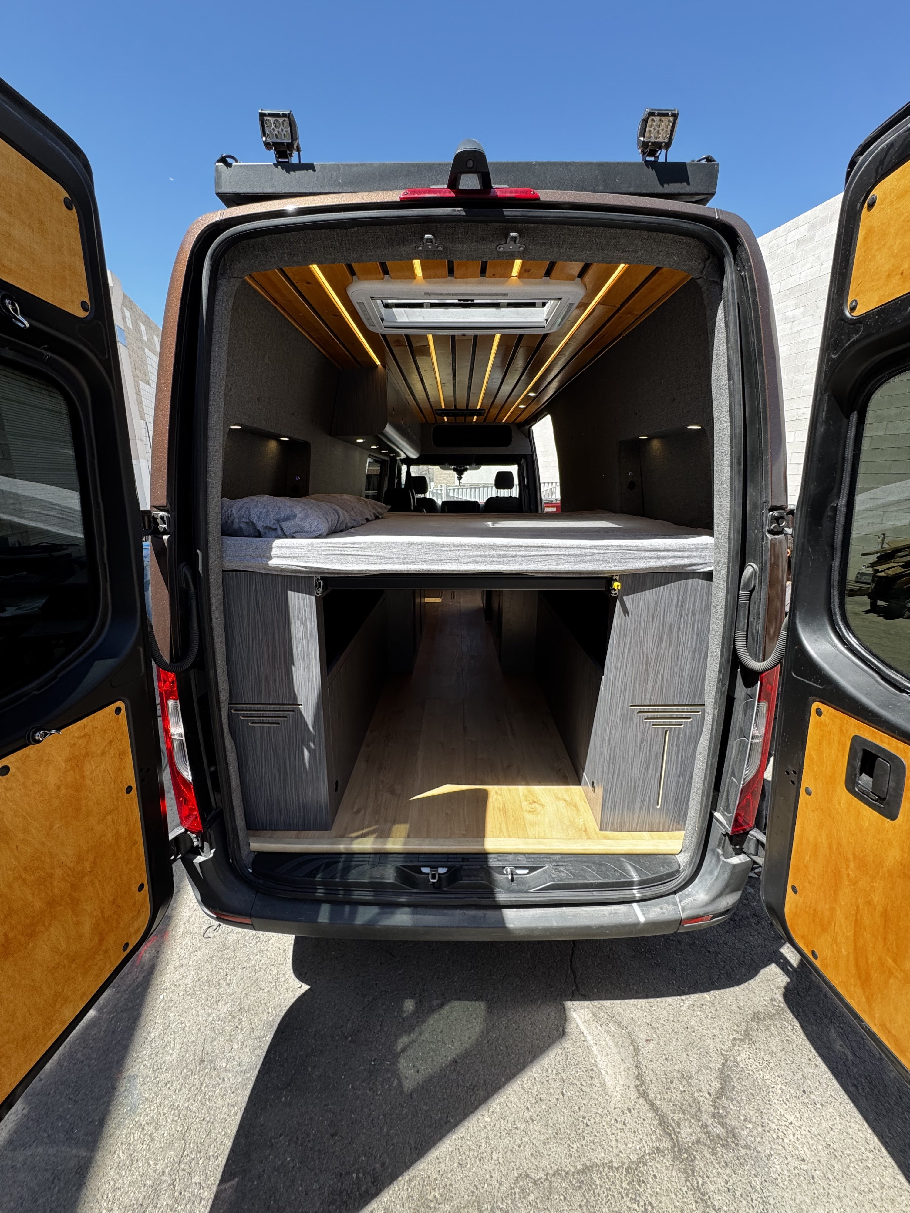 Modern van conversion RV rental: rear view revealing a comfortable bed, under-bed storage, and wood-slat ceiling with lights.. Mercedes-Benz Sprinter 2023