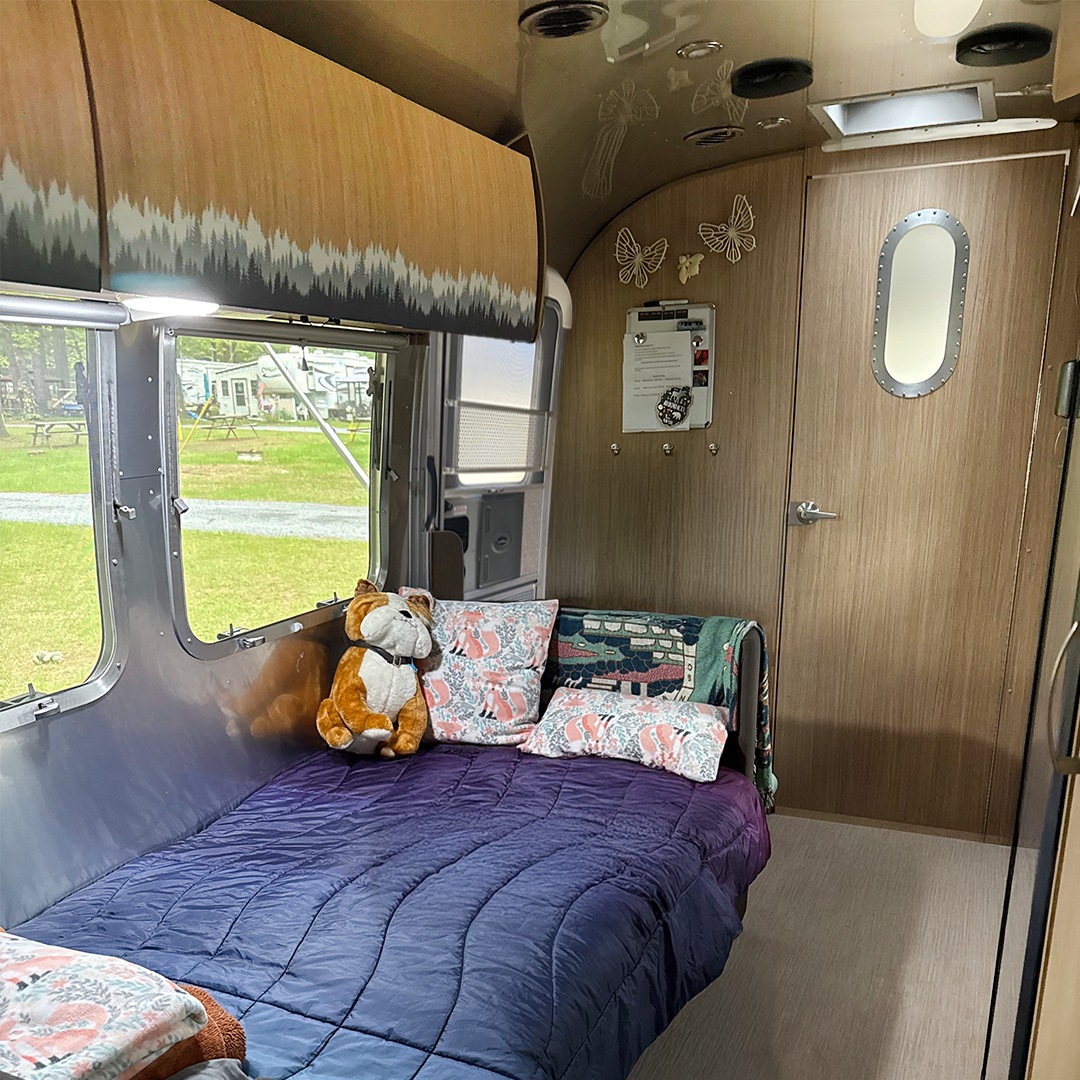 Inside a comfy rv rental bedroom: purple bed, forest-themed cabinets, window view of camp.. Airstream Flying Cloud 2018