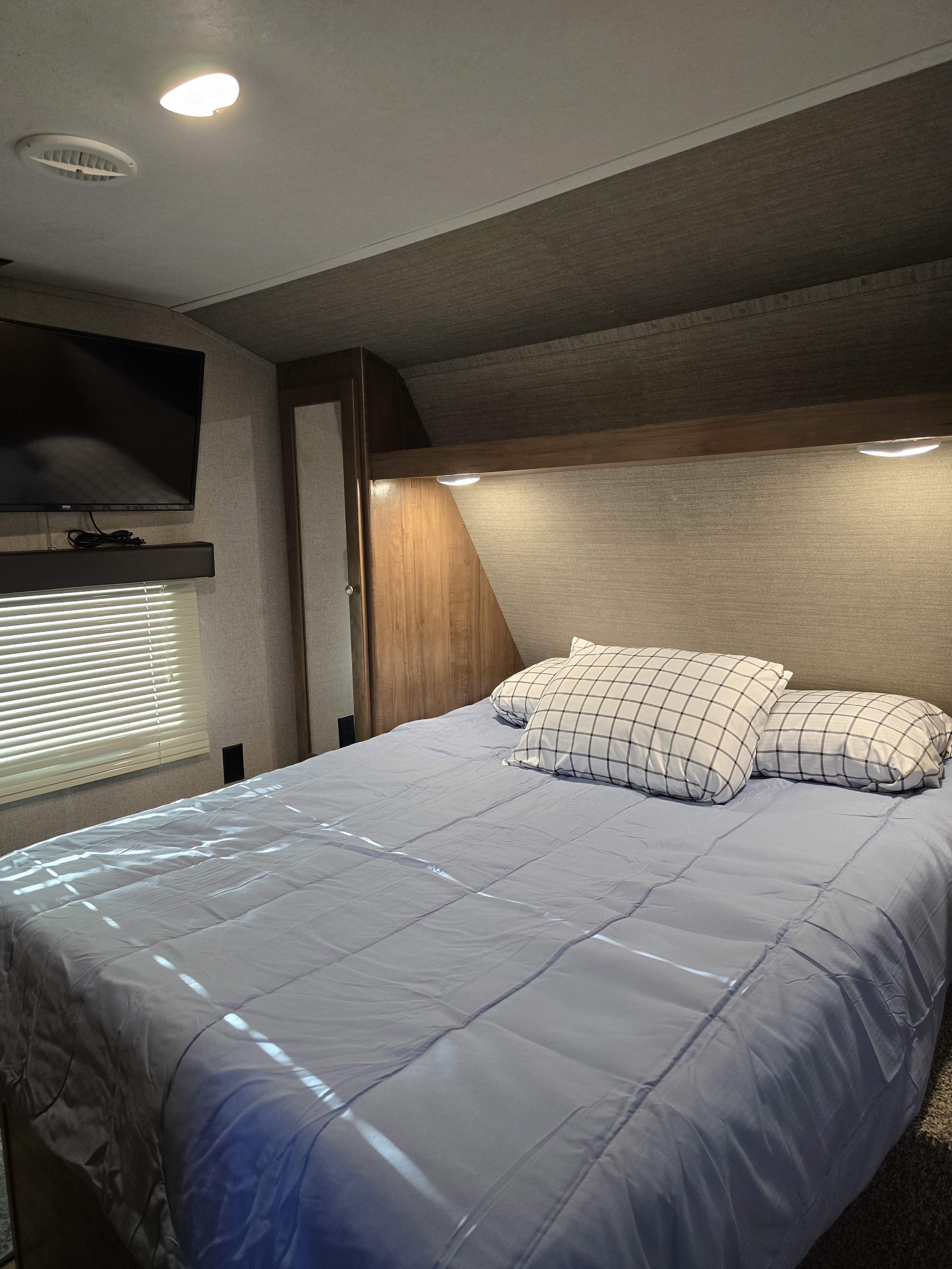 Cozy RV rental bedroom with blue comforter, plaid pillows, TV, and bright interior lights, ideal for travel.. Aspen Trail Aspen Trail Trailer 2018