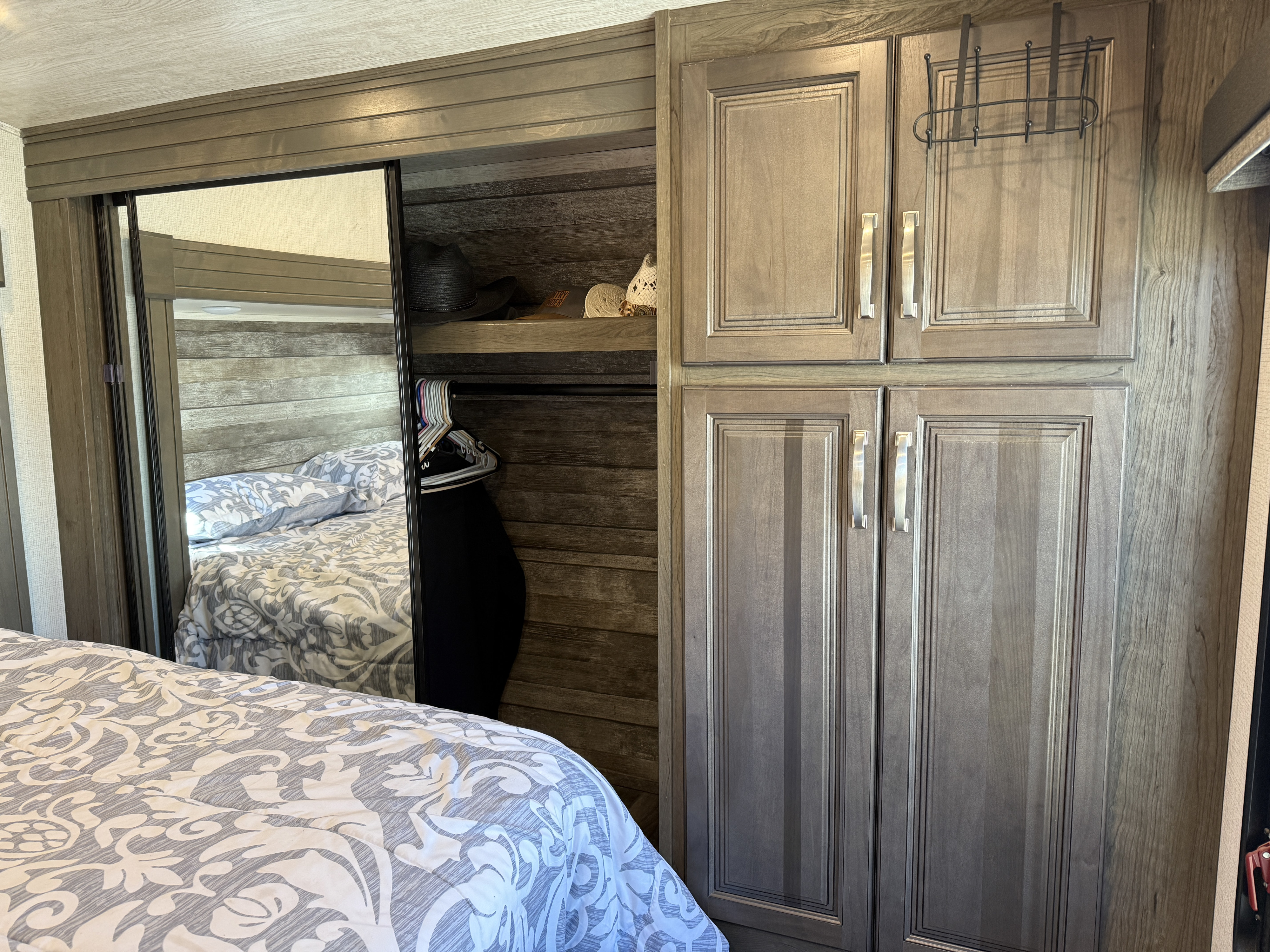 RV rental bedroom with a mirrored closet, revealing a bed with patterned linens, alongside hanging clothes, hats, and built-in cabinets.. Keystone RV Cougar 2022