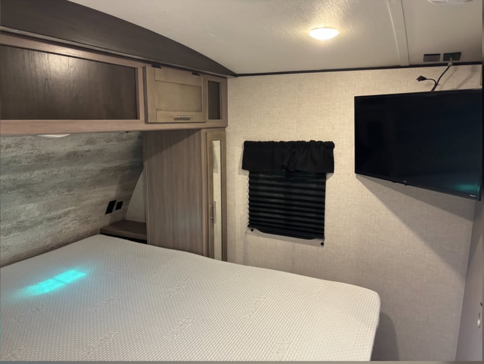 Compact RV rental bedroom featuring a white bed, overhead cabinets, window with blinds, and wall-mounted TV.. Crossroads RV Sunset Trail Super Lite 2022