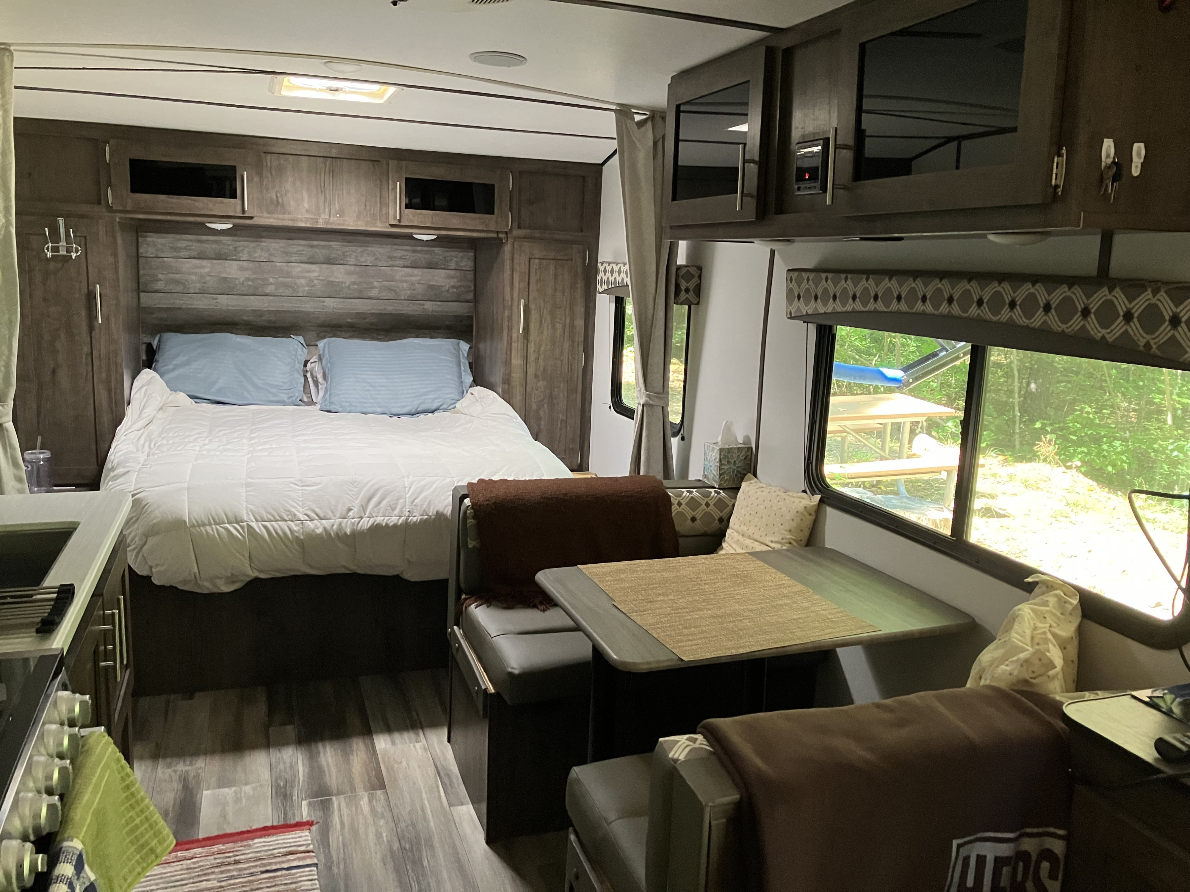 Inside a comfy RV rental: bedroom with a bed, a dining booth, and a kitchen area. Windows offer views of a serene outdoor campsite.. Riverside RV Retro 2021