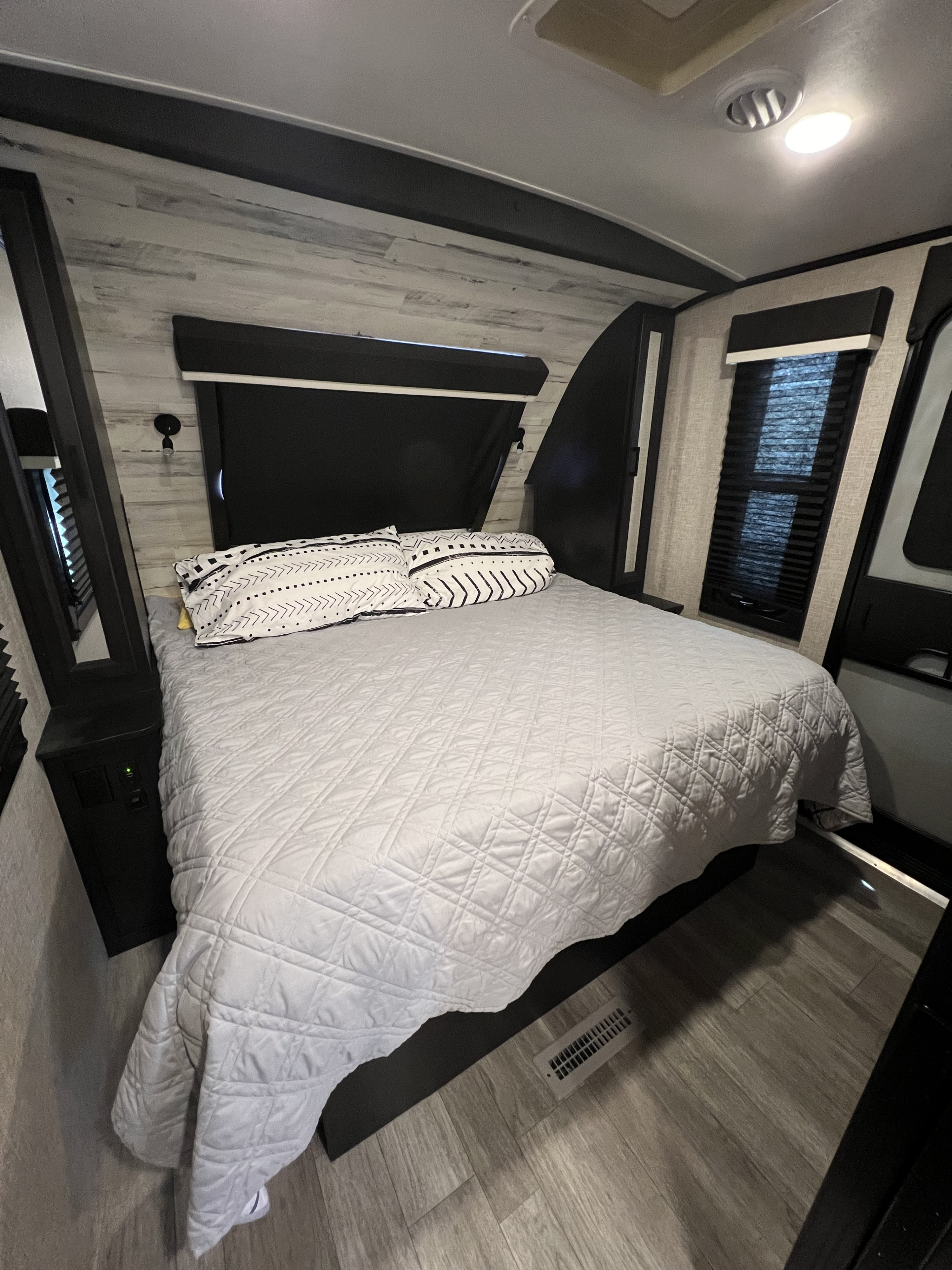This RV rental bedroom features a comfy queen bed with patterned pillows, wood-look walls, and bright overhead lighting.. Jayco White Hawk 2022